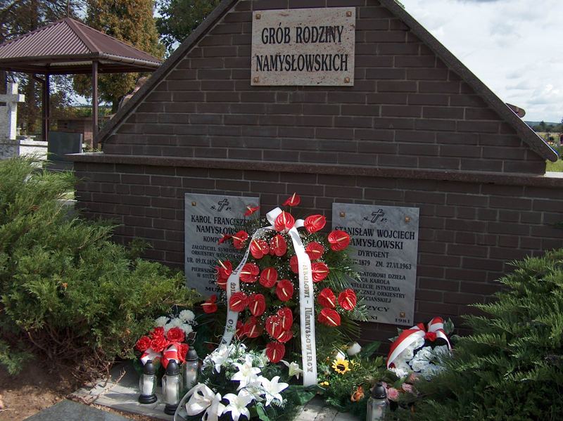 Zdjęcie grobu rodziny Namysłowskich zbudowanego z ciemnej cegły. Na grobie są 3 wieńce i 4 znicze. Po bokach widać zielone krzewy. 