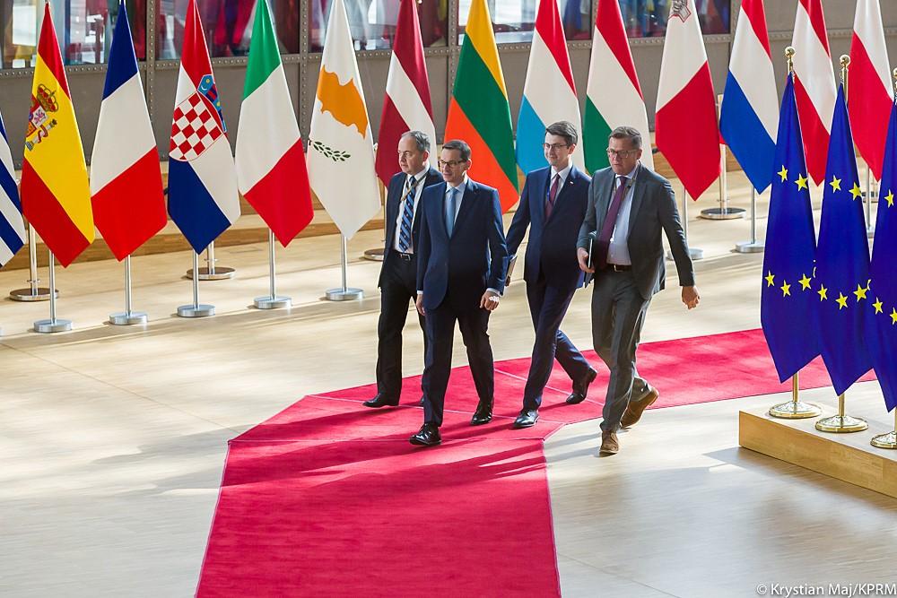 Minister Konrad Szymański, premier Mateusz Morawiecki oraz minister Piotr Muller idą po czerwonym dywanie na tle flag.