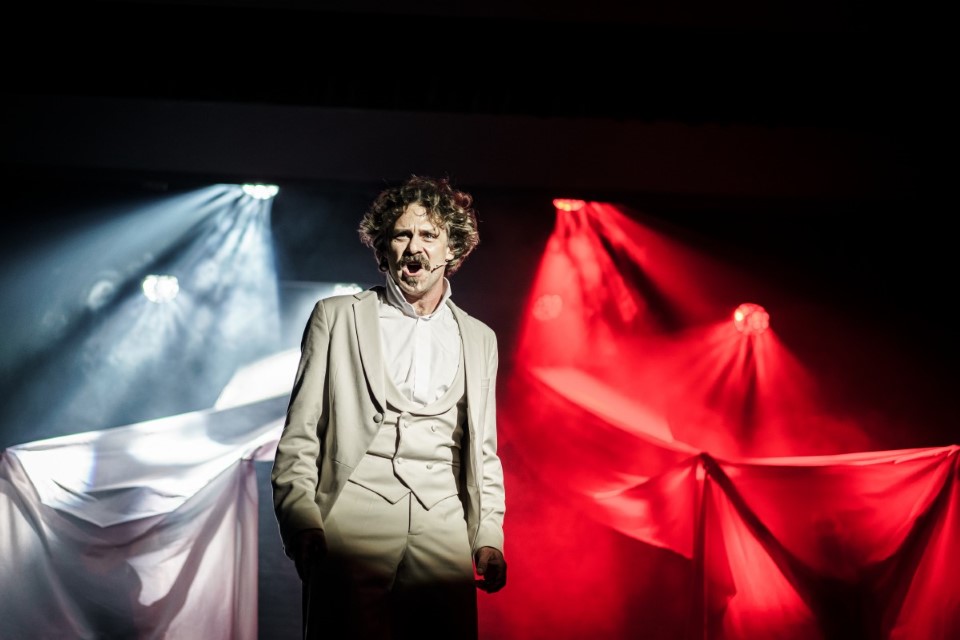 Zdjęcie kolorowe. Aktor w stroju stylizowanym na frak z XIX wieku na scenie. Śpiewa. W tle biało czerwona flaga.