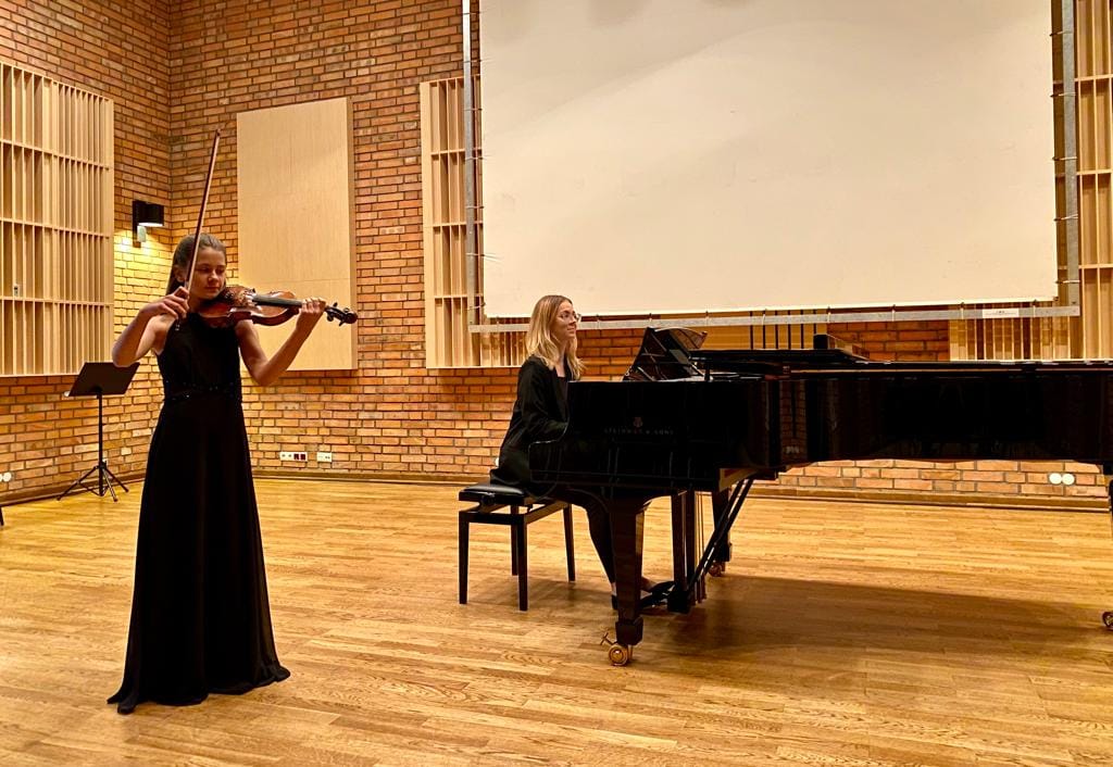 Na zdjęciu znajduje się uczennica Agata Kostanowicz oraz nauczycielka Monika Duda. Uczennica gra na skrzypcach, ma na sobie długą, czarną sukienkę. Nauczycielka akompaniuje przy czarnym fortepianie. 