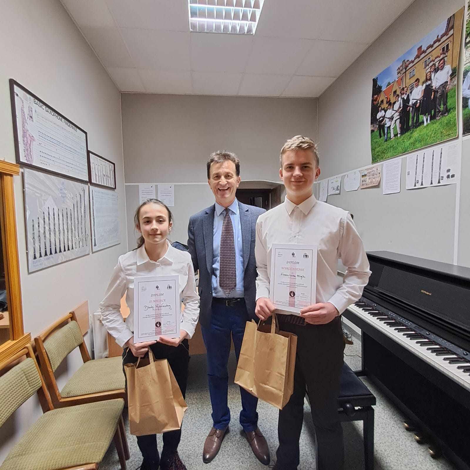 Zdjęcie przedstawia nauczyciela i dwójkę uczniów trzymających dyplomy i nagrody. 