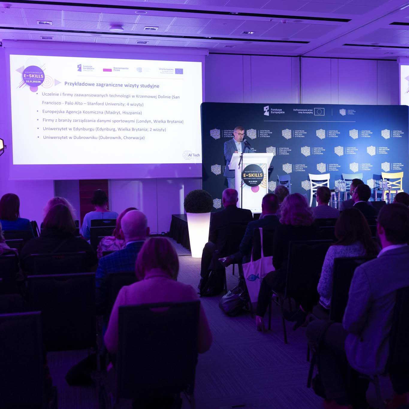 Przy mównicy stoi profesor Jacek Marciniak. Wzrok kieruje w dół. Mówi do mikrofonu. Zdjęciem objęta jest sala konferencyjna, w tle, na ekranie wyświetlona jest prezentacja dotycząca projektu AI Tech.
