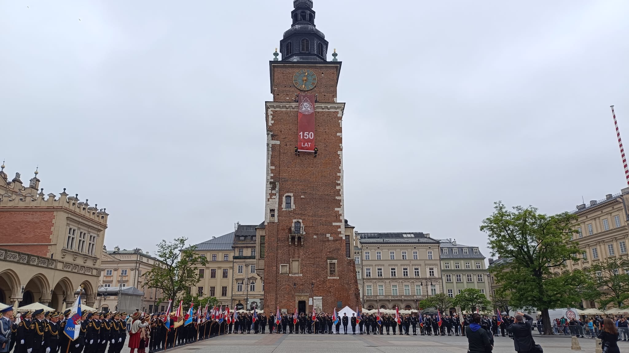 Rynek Krakowa, kamienice i wieża ratuszowa na której wisi flaga jubileuszowa z napisem 150 lat Krakowskiej Straży Pożarnej, na placu stoją uczestnicy obchodów