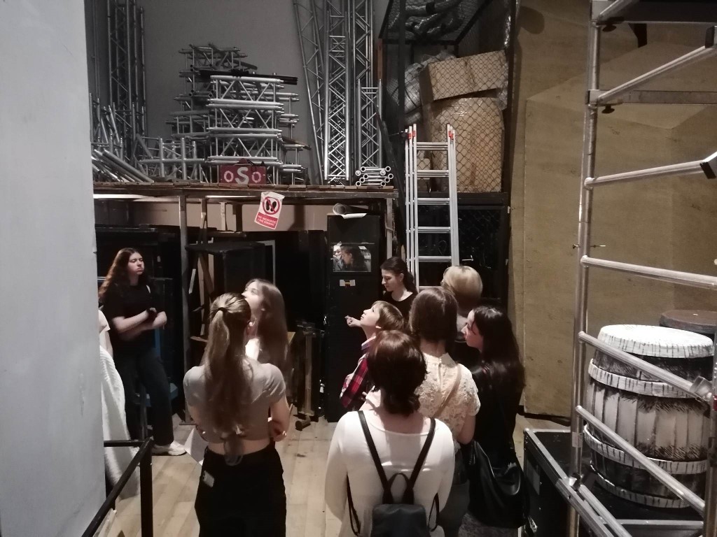 Zdjęcie kolorowe. Grupa uczniów na zapleczu sceny teatru. Wokół elementy dekoracji przedstawień teatralnych.