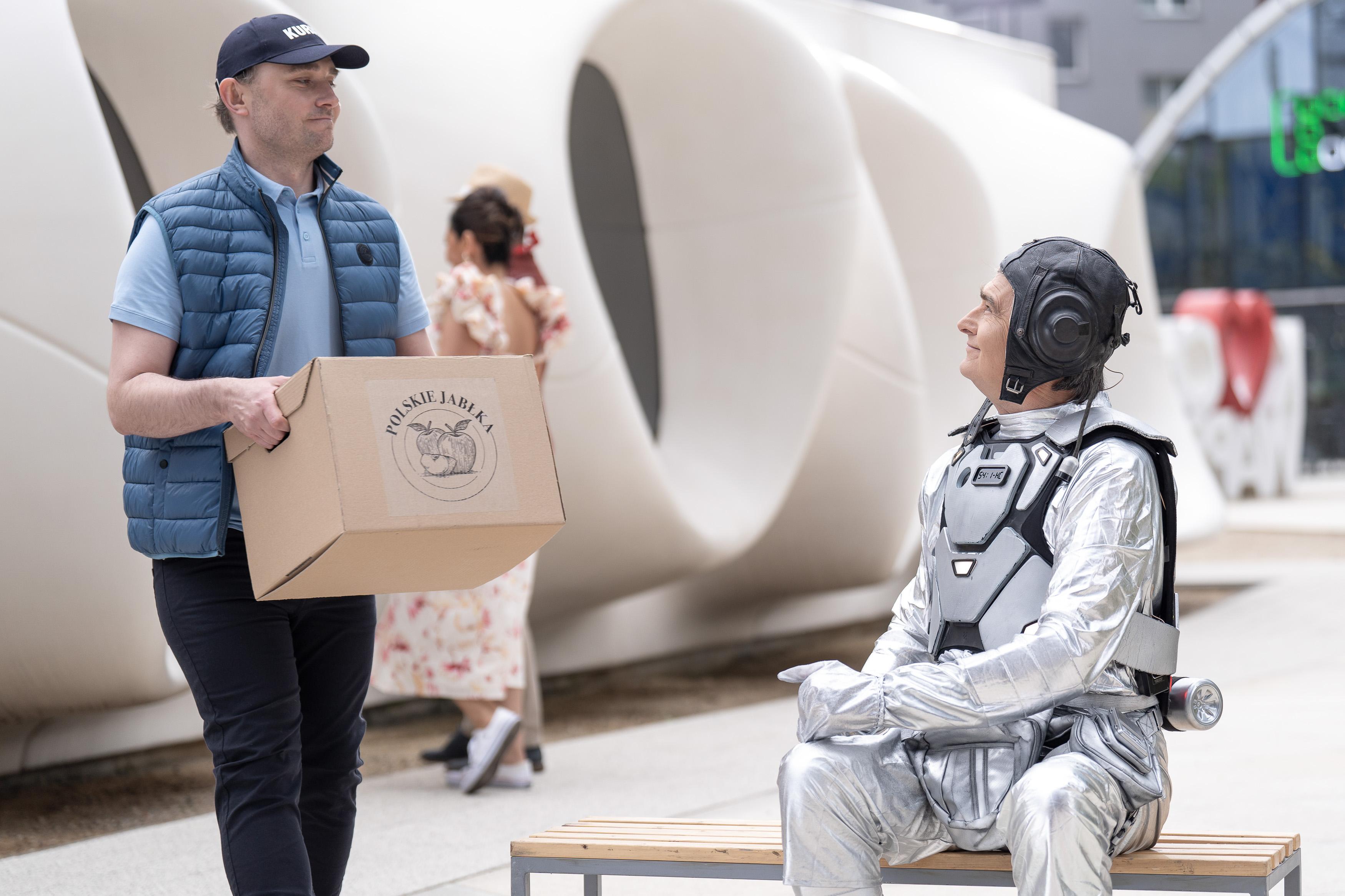 Uśmiechnięty mężczyzna w stroju astronauty siedzi na ławce i patrzy na mężczyznę, który trzyma kartonowe pudło z napisem "polskie jabłka"