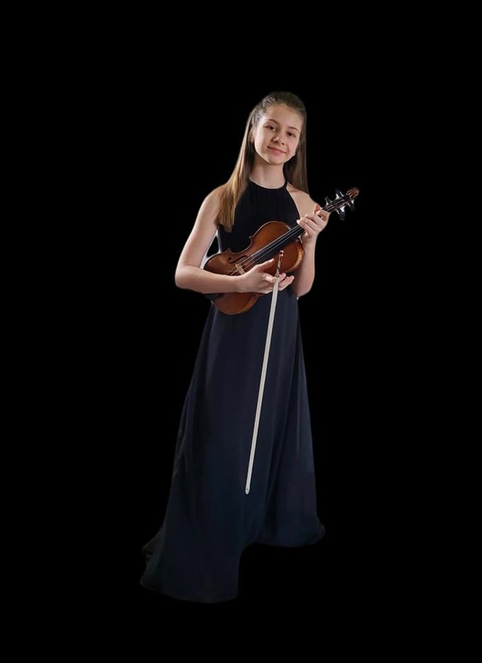 Na zdjęciu widoczna jest uczennica Agata Kostanowicz. Jest ubrana w czarną suknię, w rękach trzyma skrzypce i smyczek. Zdjęcie ma czarne tło.