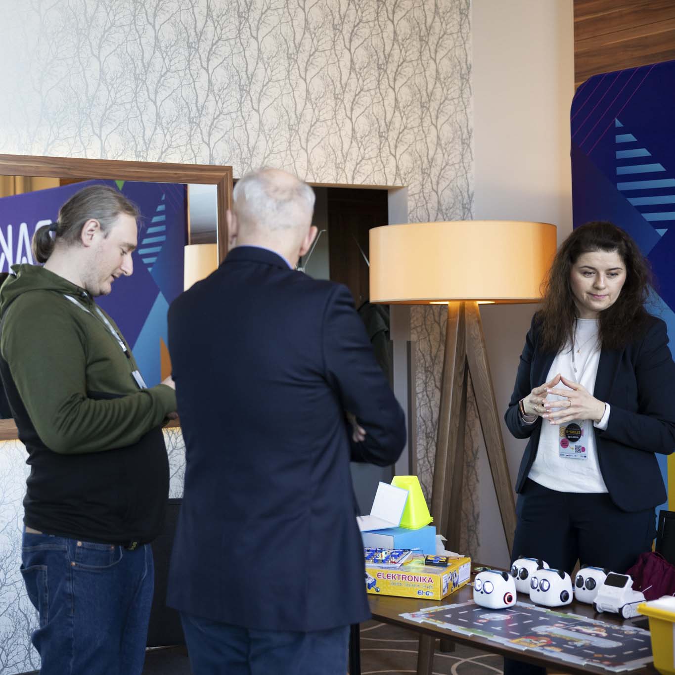 Dwóch mężczyzn stoi przy stoisku Pracowni Aktywnego Korzystania z Technologii, naprzeciwko stoi kobieta patrząc w lewo.