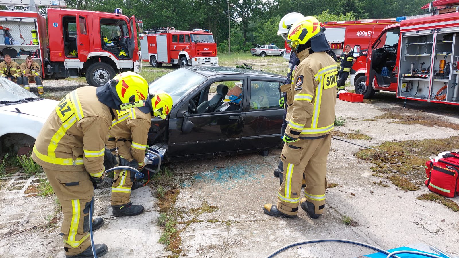 Strażacy rozcinają nożycami hydraulicznymi samochód osobowy, w tle samochody ratowniczo - gaśnicze 