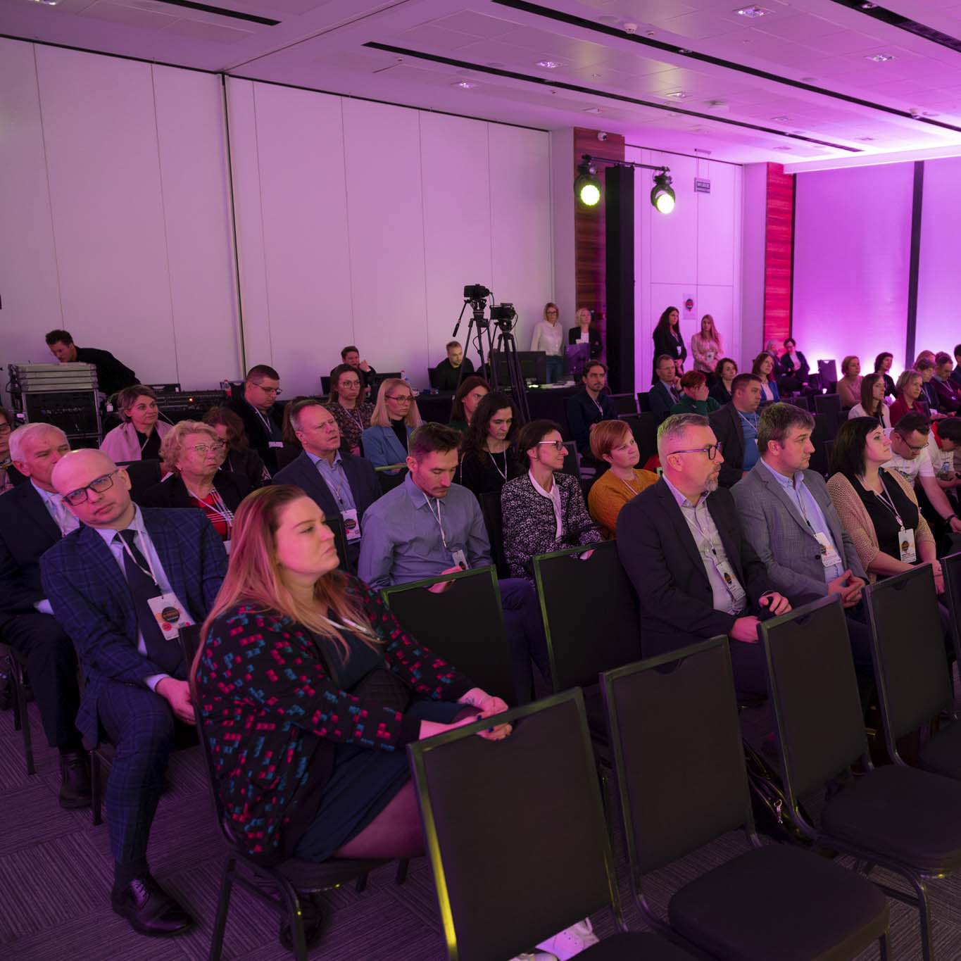 Na krzesłach siedzą uczestnicy i uczestniczki konferencji. Wzrok kierują w stronę sceny.