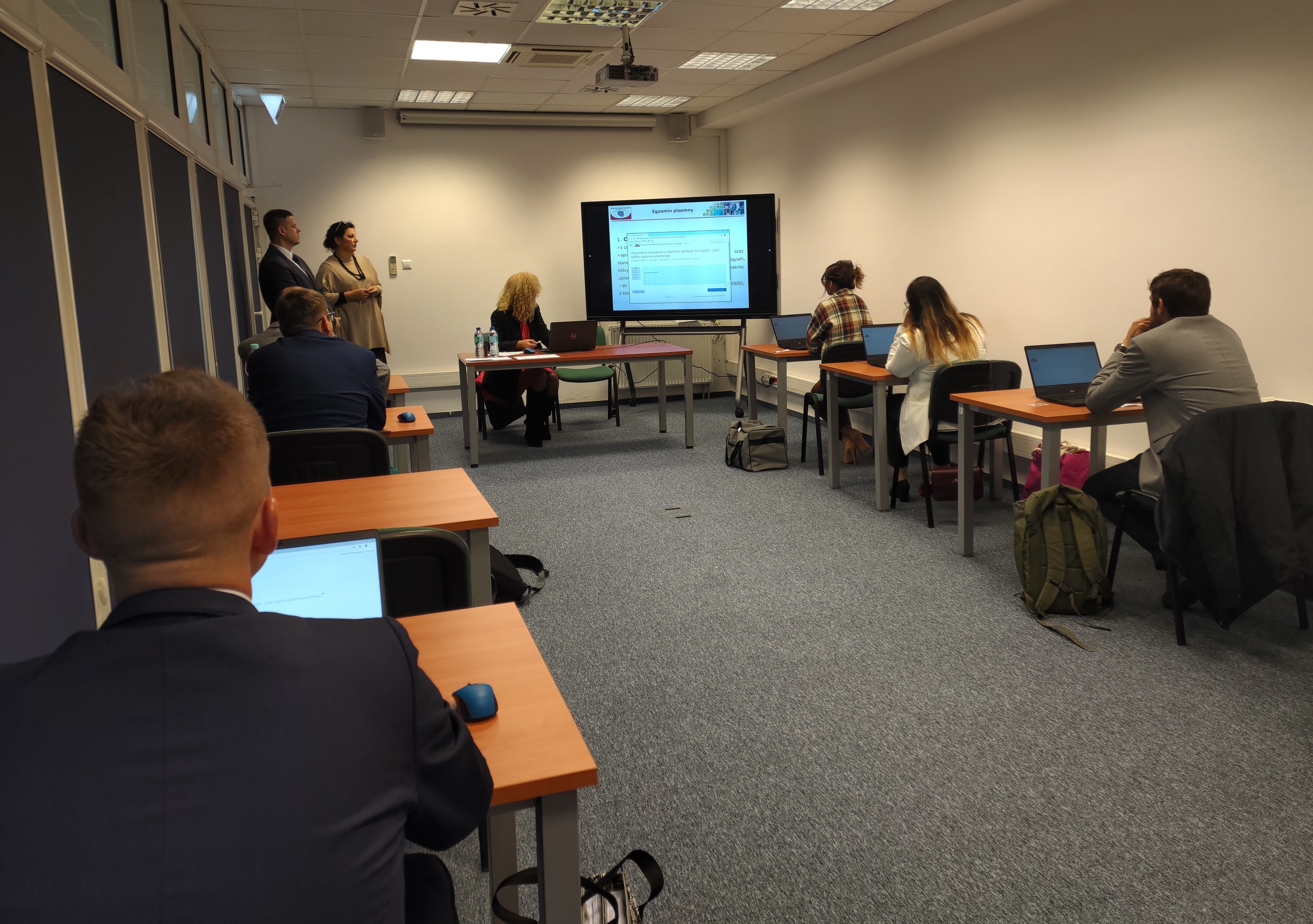 Zdjęcie przedstawia salę egzaminacyjną z z członkami Komisji egzaminacyjnej i osobami przystępującymi do egzaminu siedzącymi pojedynczo przed laptopami.