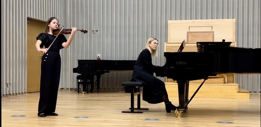 Zdjęcie zostało wykonane w sali koncertowej. Widoczne są na nim uczennica grająca na skrzypcach oraz akompaniująca jej na fortepianie nauczycielka.