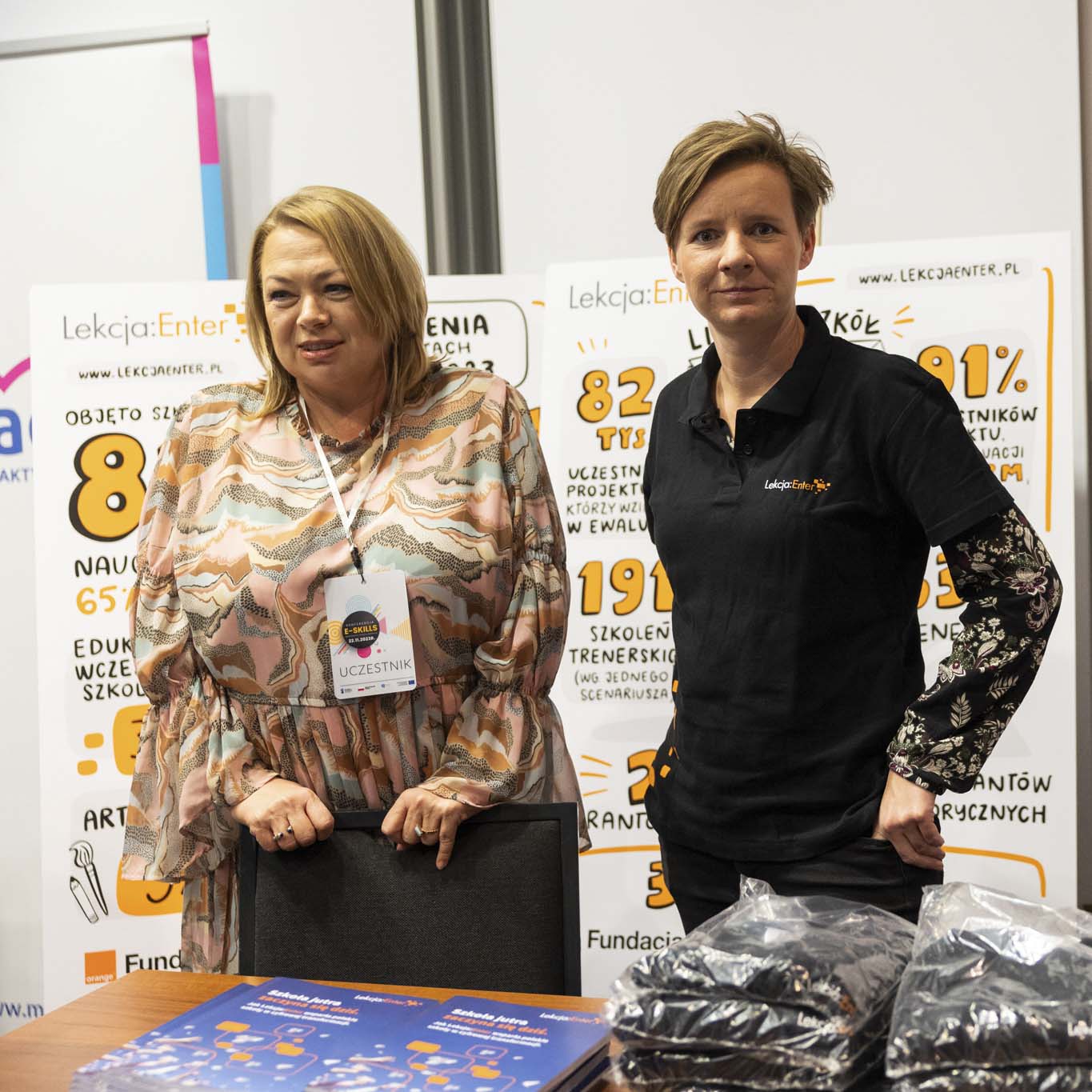 Dwie kobiety stoją na tle plakatu projektu Lekcja Enter. Jedna z nich patrzy w obiektyw, druga spogląda w prawą stronę. 