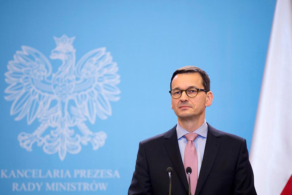 Premier Mareusz Morawiecki podczas konferencji prasowej na tle niebieskiej ścianki i godła.
