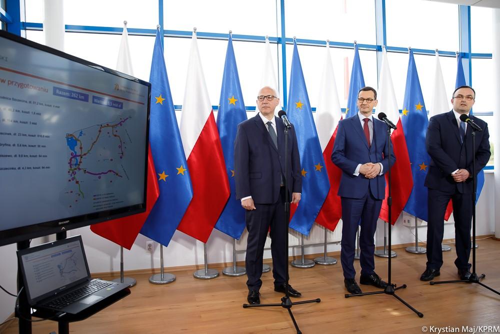 Premier Mateusz Morawiecki, minister Joachim Brudziński i wojewoda zachodniopomorski stoją przy mikrofonach i patrzą na prezentację.