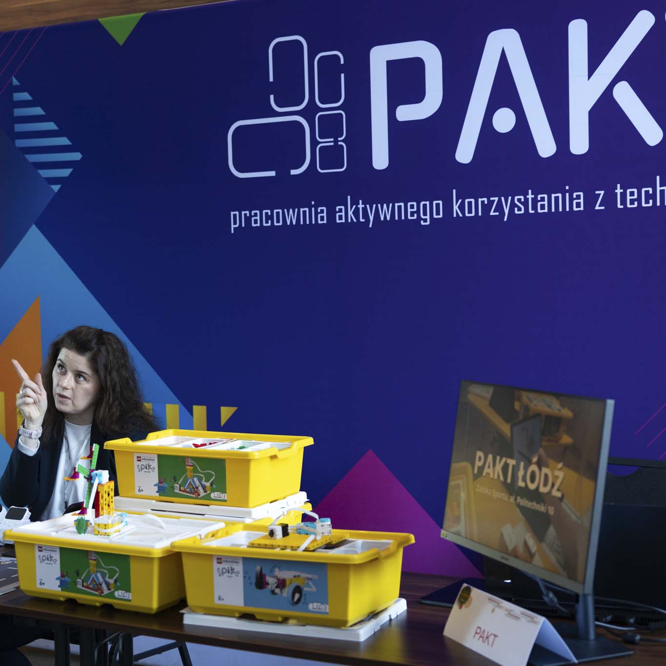 Przy stoisku Pracowni Aktywnego Korzystania z Technologii stoi mężczyzna, siedząca obok kobieta palcem wskazującym pokazuje na niego. Na pierwszem planie stoją klocki, roboty.