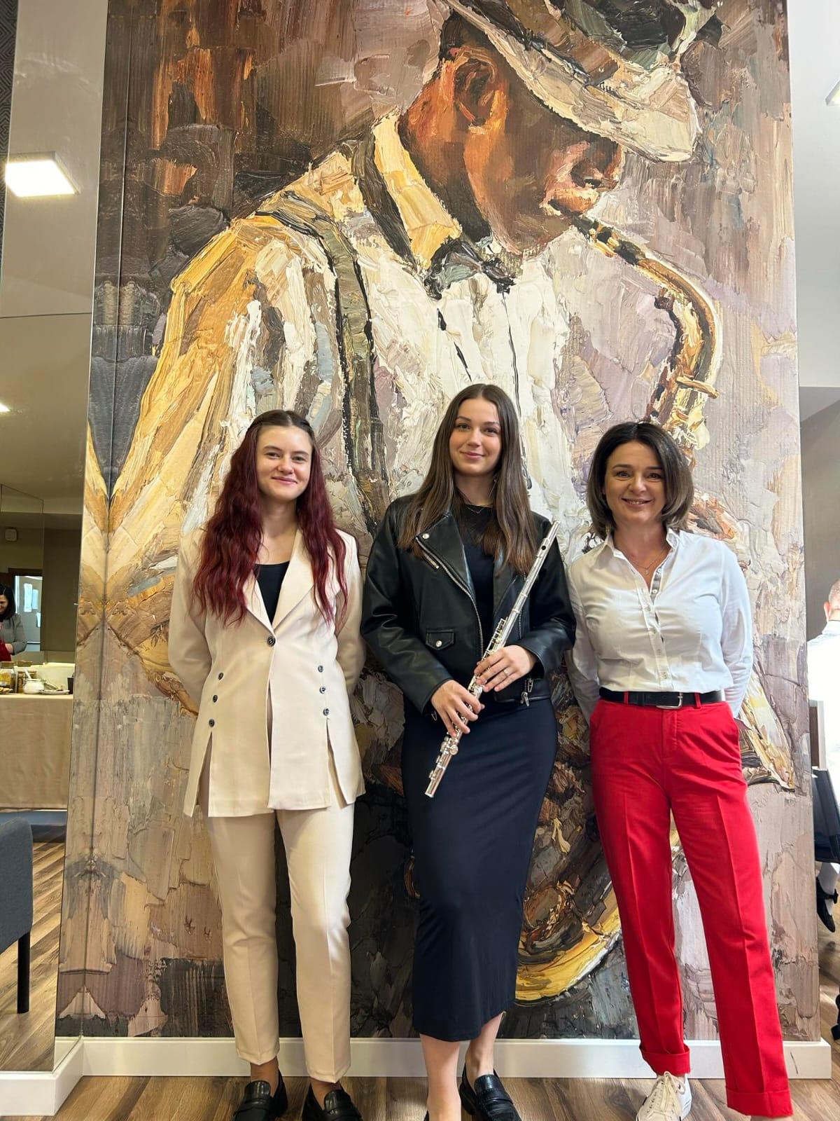 Po środku uczennica trzymająca flet, a po jej bokach dwie nauczycielki. W tle widoczny obraz przedstawiający mężczyznę grającego na saksofonie.