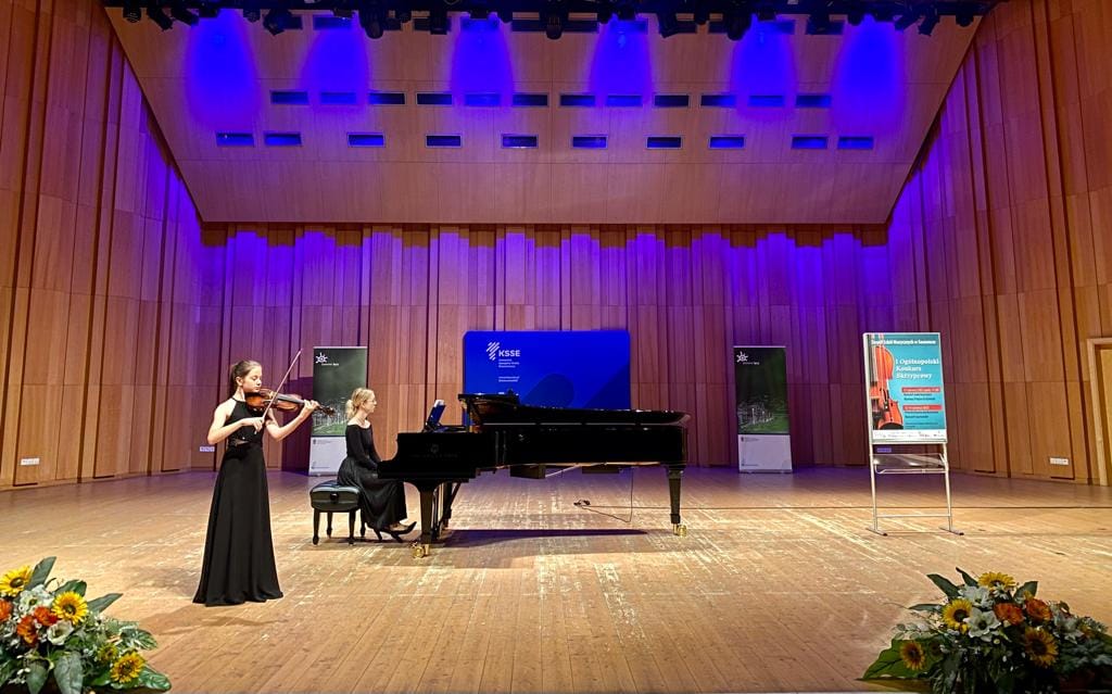 Zdjęcie przedstawia prezentację konkursową w sali koncertowej. Na zdjęciu widoczna dziewczynką w czarnej, długiej sukni, która gra na skrzypcach. Towarzyszy jej akompaniatorka, również w czarnej długiej sukni grająca na czarnym fortepianie. W tle banery reklamowe, konkursowe, z przodu kwiaty w formie dekoracji. 