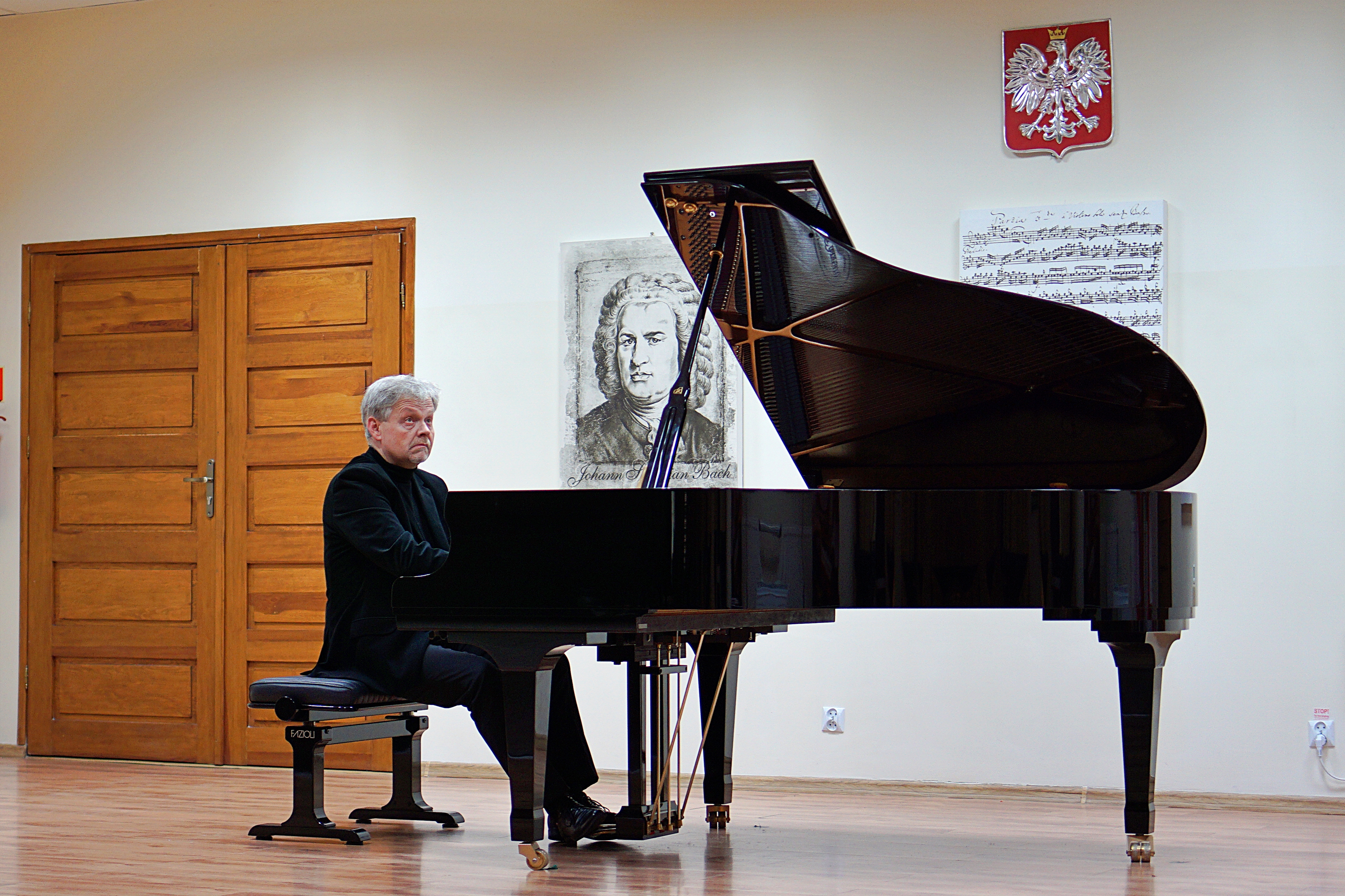Zdjęcie przedstawia scenę sali koncertowej Państwowej Szkoły Muzycznej w Grajewie. Na scenie widoczny jest mężczyzna ubrany na czarno i grający na fortepianie. W tle widać godło Polski, brązowe drzwi oraz grafiki przedstawiające nuty i portret J. S. Bacha. 