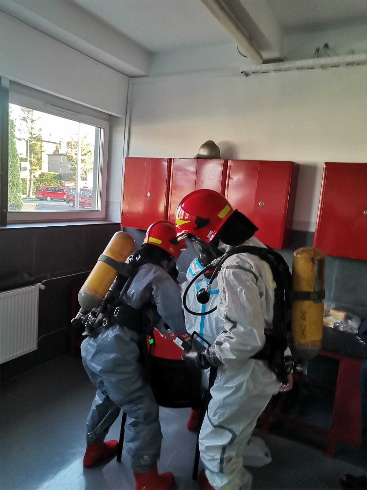 trzech funkcjonariuszy ubranych w kombinezony ochronne, czerwone hełmy oraz aparaty powietrzne podczas ćwiczeń z zakresu ratownictwa chemicznego i ekologicznego 