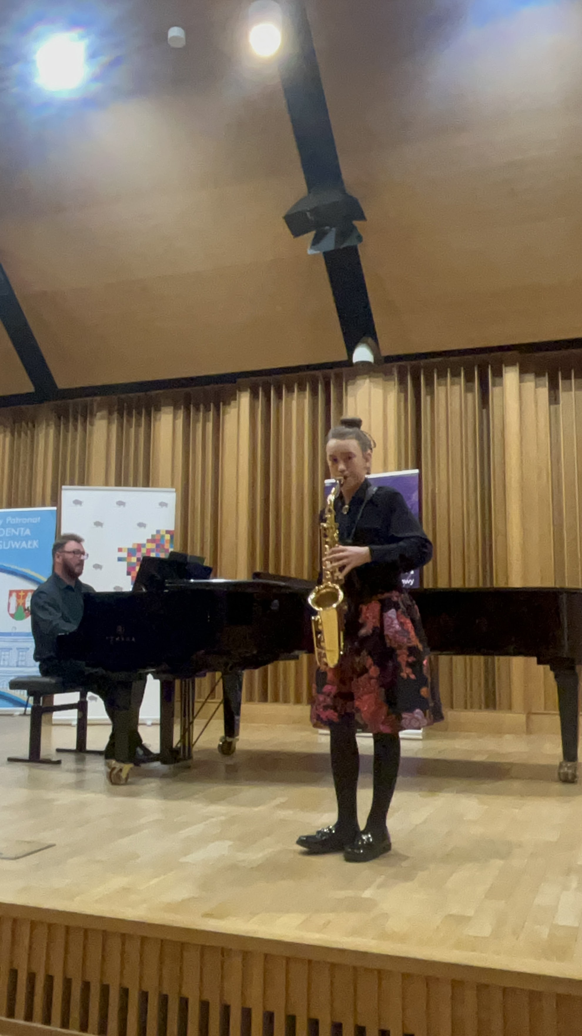 Zdjęcie przedstawia dziewczynkę grającą na złotym saksofonie. Jest ubrana we wzorzystą sukienkę. W dalszym planie widać akompaniatora ubranego na czarno grającego na czarnym fortepianie. Oboje są na scenie.