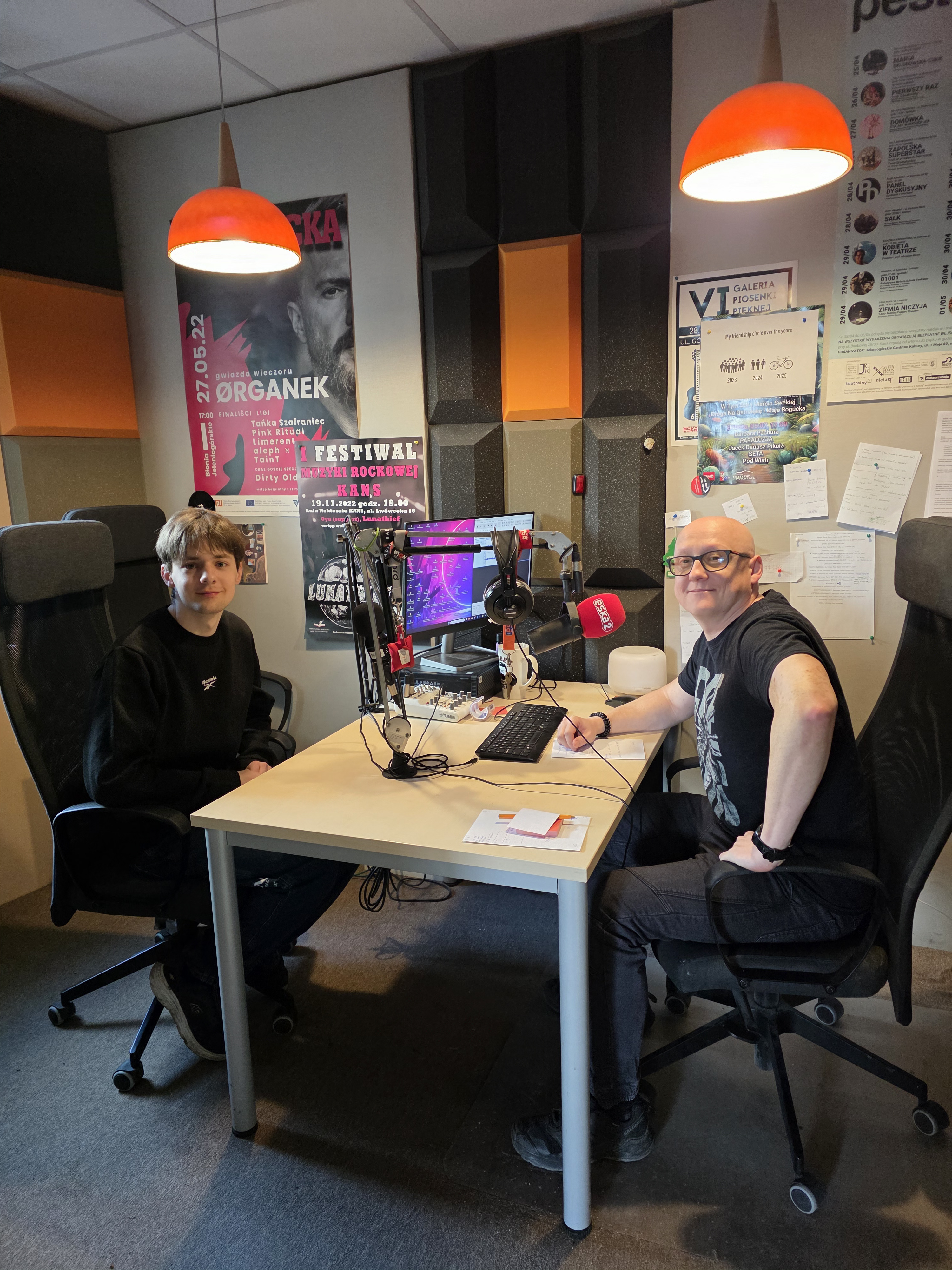 Zdjęcie przedstawia wnętrze nowoczesnego studia radiowego, w którym panuje profesjonalna, a jednocześnie swobodna atmosfera. W centralnej części kadru, przy jasnym prostokątnym biurku, siedzą naprzeciw siebie dwaj mężczyźni. Po lewej stronie znajduje się młodsza osoba w czarnej bluzie, patrząca spokojnie w stronę obiektywu, natomiast po prawej siedzi uśmiechnięty mężczyzna w okularach i ciemnym t-shircie, który trzyma długopis nad notatkami. Przestrzeń roboczą wypełnia specjalistyczny sprzęt: na biurku stoi monitor z fioletowym interfejsem programu muzycznego, klawiatura oraz mikser. Przed prowadzącym zamontowano profesjonalny mikrofon na ruchomym ramieniu, osłonięty czerwoną gąbką z logo „eska”. Tło tworzą ściany wyłożone szarymi i pomarańczowymi panelami akustycznymi o geometrycznych kształtach, na których gęsto rozwieszono plakaty koncertowe, w tym afisz artysty o pseudonimie Organek oraz ogłoszenia o festiwalach rockowych. Całość kompozycji dopełniają dwie wiszące lampy o jaskrawopomarańczowych kloszach, które wypełniają studio ciepłym, kameralnym światłem.