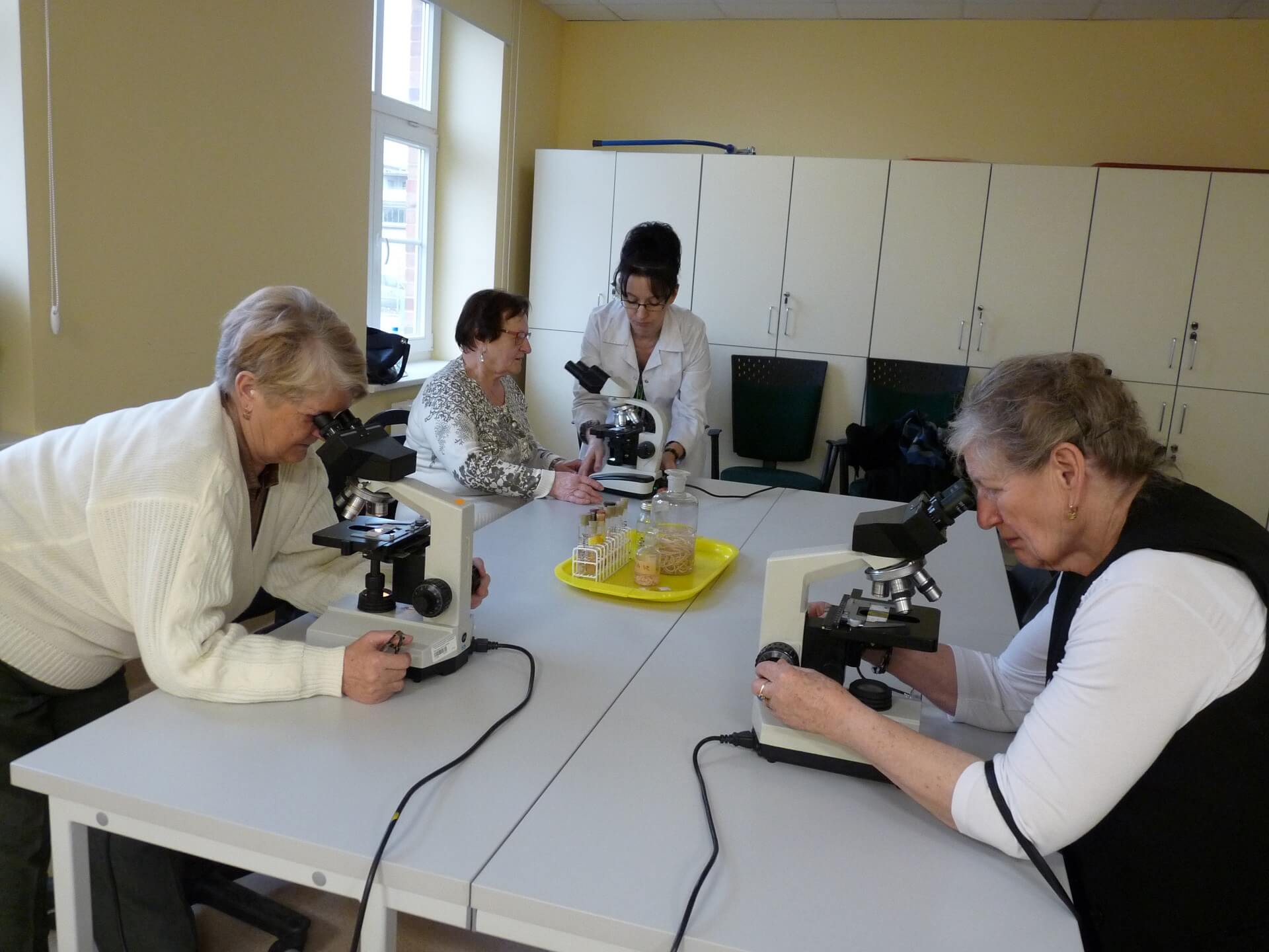 Zajęcia na Wydziale Biotechnologii i Hodowli Zwierząt z udziałem grupy seniorów. Trzy panie prowadzą obserwacje przy mikroskopach pod okiem prowadzącej ubranej w biały kitel