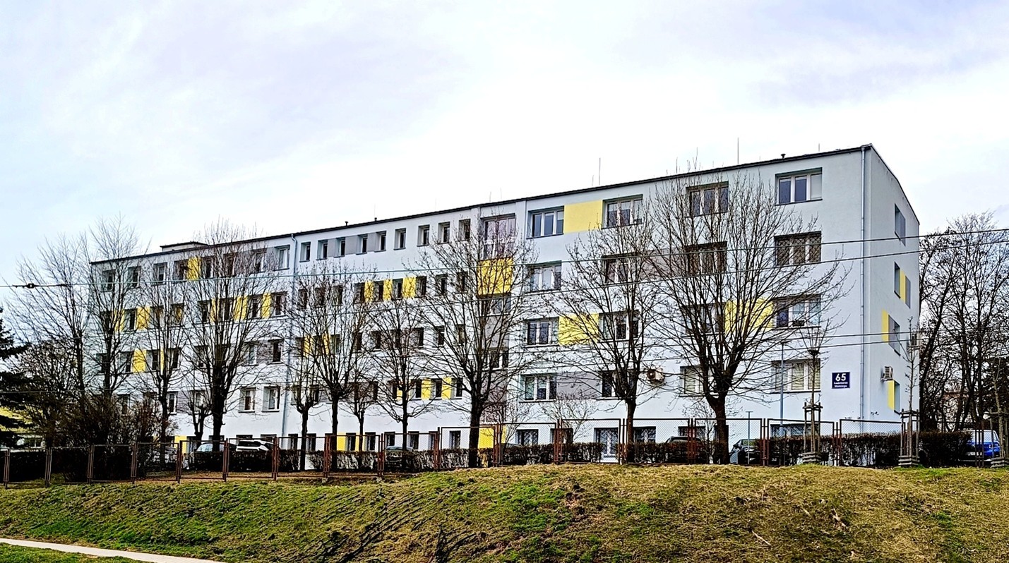 czteropiętrowy budynek prostokątny, elewacja szara z elementami żółtego, przed nim trawnik