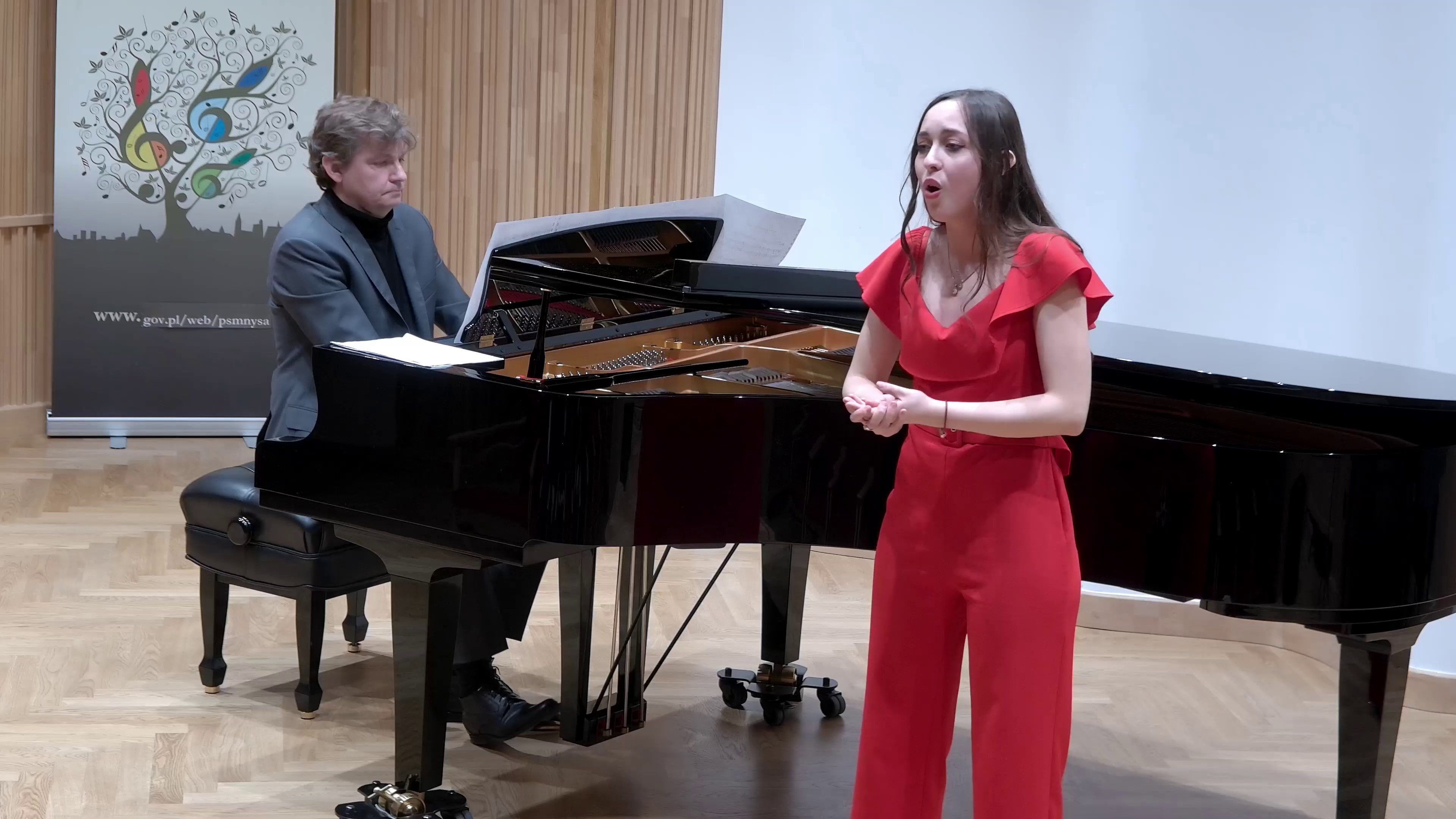 Zdjęcie. Na estradzie występuje ubrana w czerwony strój śpiewaczka z towarzyszeniem pianisty, który gra na dużym czarnym fortepianie. W głębi z lewej strony widać baner PSM Nysa.