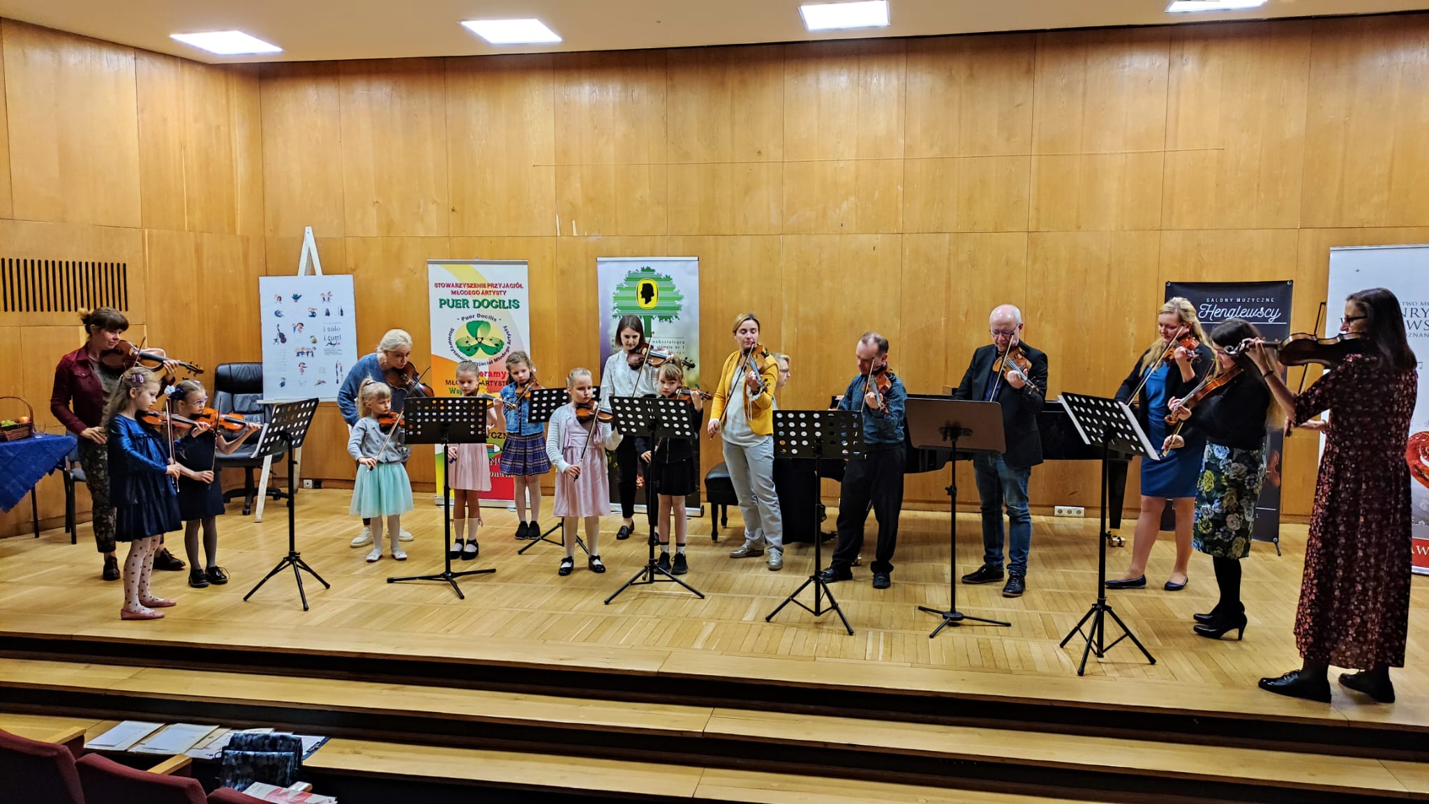 Zdjęcie przedstawia uczniów wraz z nauczycielami, którzy grają na skrzypcach. 
