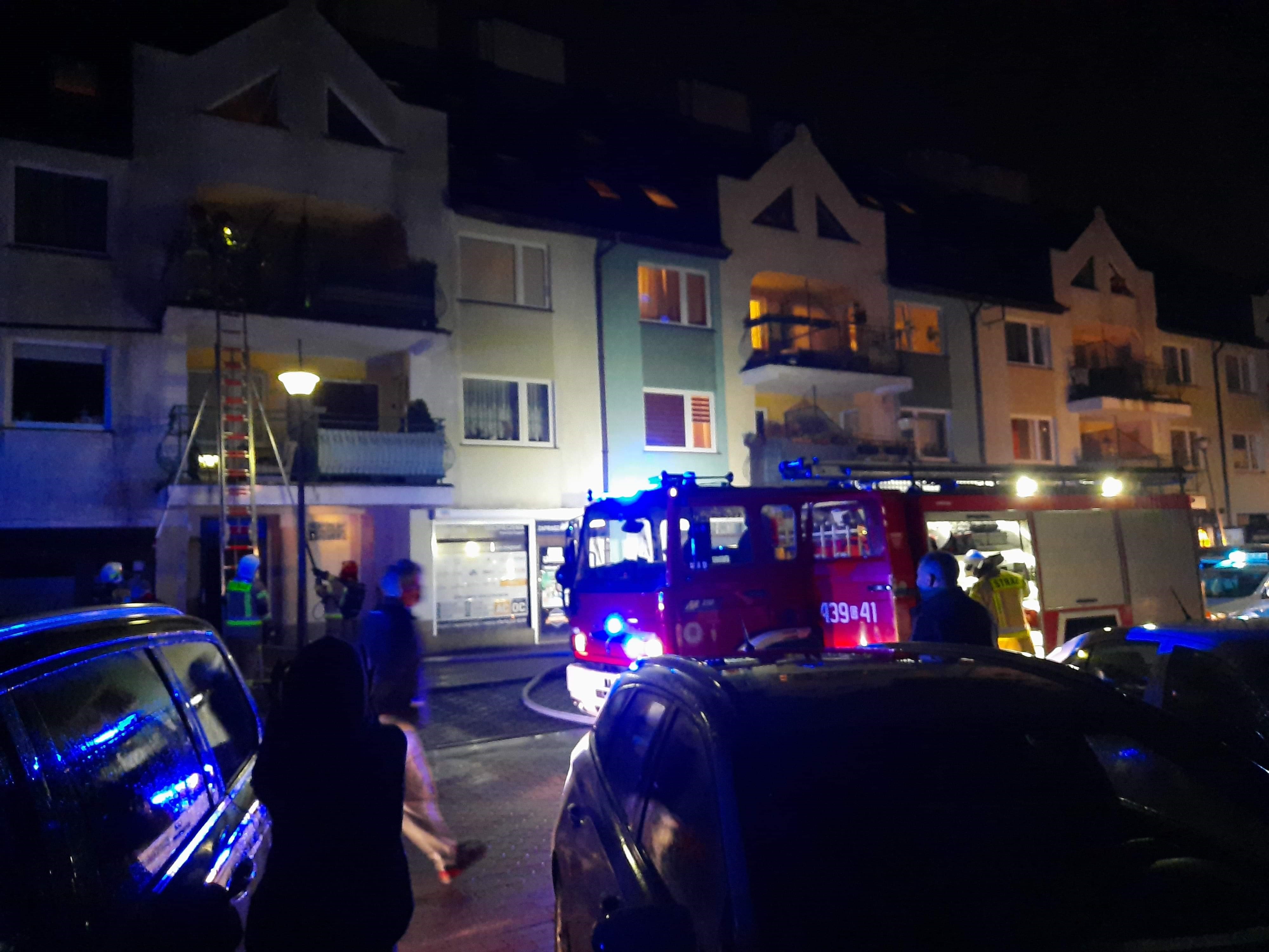 ​Strażacy przystawiają do budynku drabinę przystawną D10W. Obok stoi czerwony samochód strażacki z włączonymi, niebieskimi światłami błyskowymi. [Kliknij i przeciągnij, by przenieść.] ​