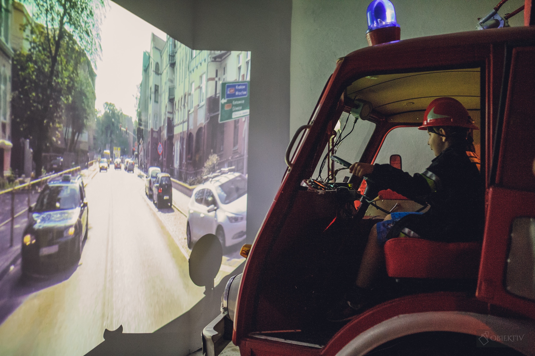 Zdjęcie przedstawia dziecko ubrane w ubranie strażaka siedzące w samochodzie pożarniczym i wyświetloną na ekranie symulację alarmowej jazdy do pożaru 