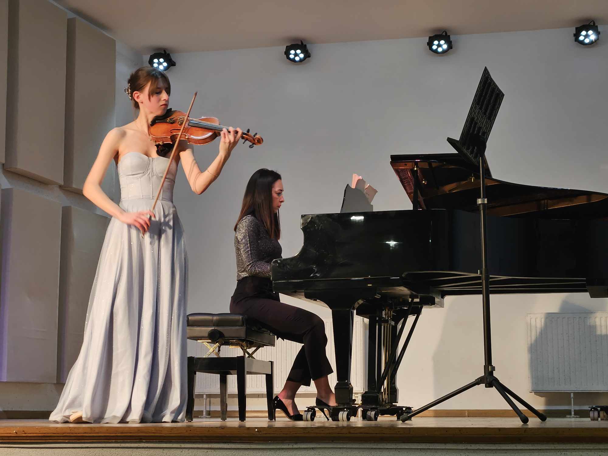 Zdjęcie przedstawia skrzypaczkę i pianistkę podczas występu na scenie. 