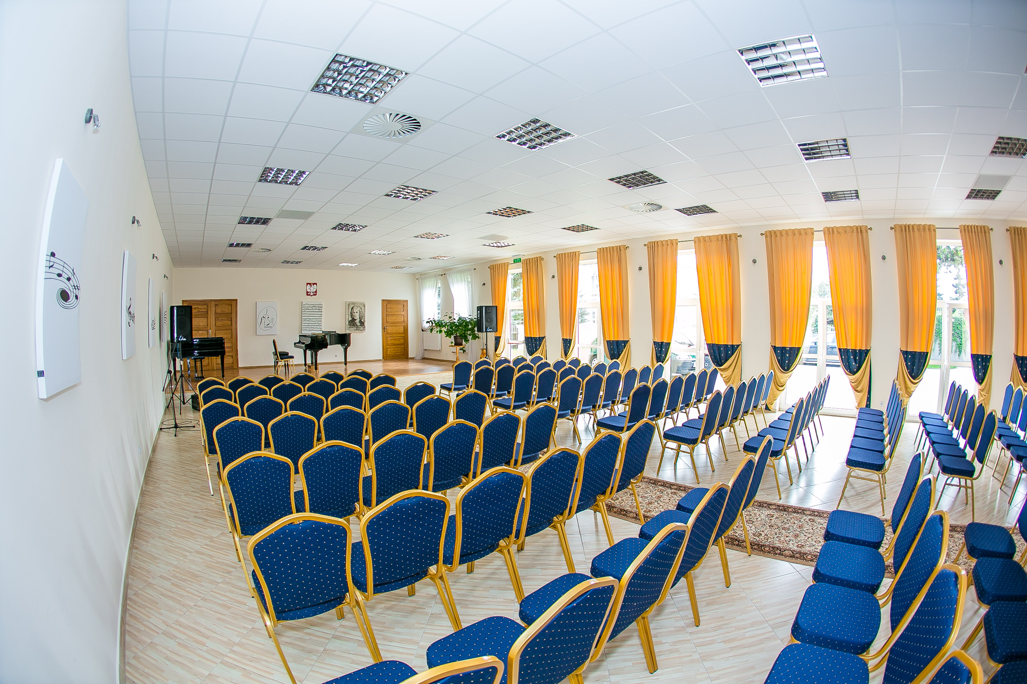Zdjęcie przedstawia wnętrze sali koncertowej, na którym widać krzesła dla publiczności w kolorze niebieskim ze złotym obramowaniem oraz scenę, na której stoi fortepian. W oknach wiszą zasłony nawiązujące kolorystycznie do krzeseł a na ścianach panele tłumiące z akcentami muzycznymi. 