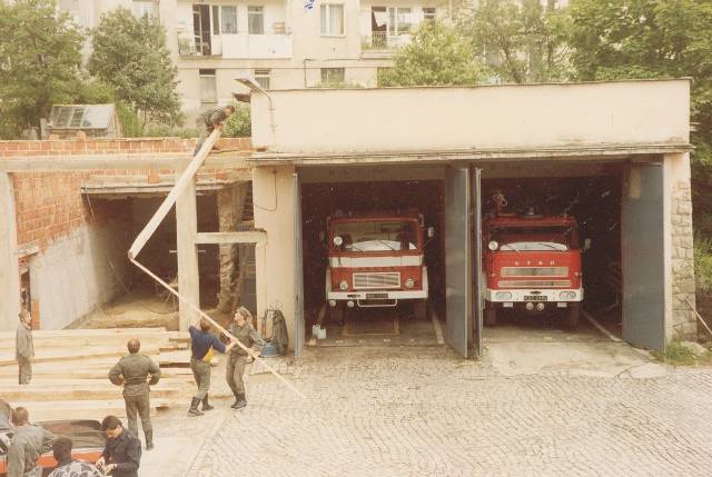 Wolnostojący garaż w JRG Krynica w rozbudowie. Na zdjęciu widoczni pracujący strażacy. W otwartych garażach widać dwa samochody pożarnicze. W tle widoczny blok mieszkalny.