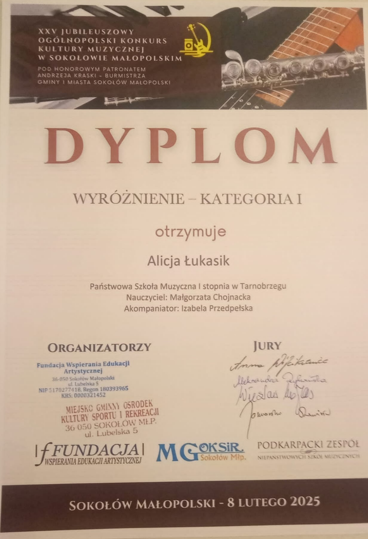 Dyplom wyróżnienia. Na górze znajduje się nazwisko patrona w brązowej ramce, a w tle zdjęcie przedstawiające fragmenty fortepianu, gitary oraz fleta poprzecznego. Po środku nazwisko otrzymującego wyróżnienie oraz nauczyciela. Na dole po lewej stronie organizatorzy, a po prawej podpisy jury. Tekst w kolorze czerwonym i czarnym.