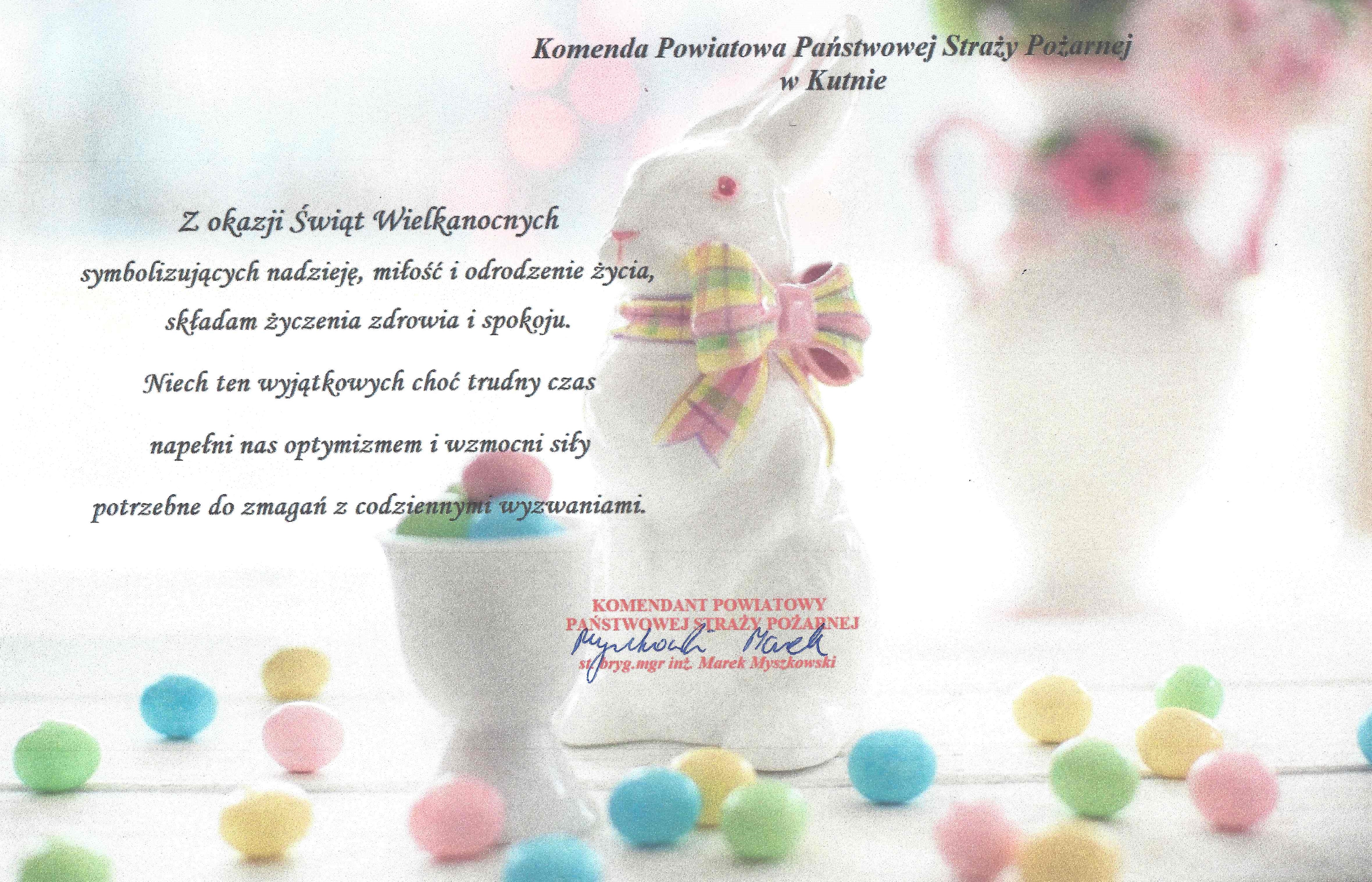 Życzenia wielkanocne na tle białego zajączka z kolorową kokardą na szyi. Dookoła zajączka leżą jajka malowane w kolorach niebieskim, żółtym, różowym i zielonym.