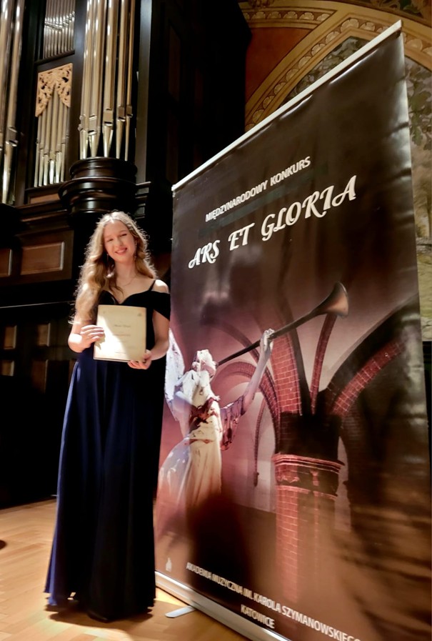Amelia Szmidt - Ars Et Gloria 2025. Uczennica pozuje do zdjęcia przy plakacie konkursu.