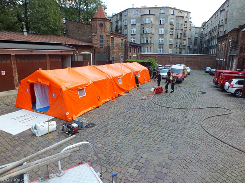 Zdjęcie przedstawia ciąg dekontaminacyjny złożony z trzech pomarańczowych namiotów na placu wewnętrznym JRG1 wraz z grupą strażaków obsługujących go