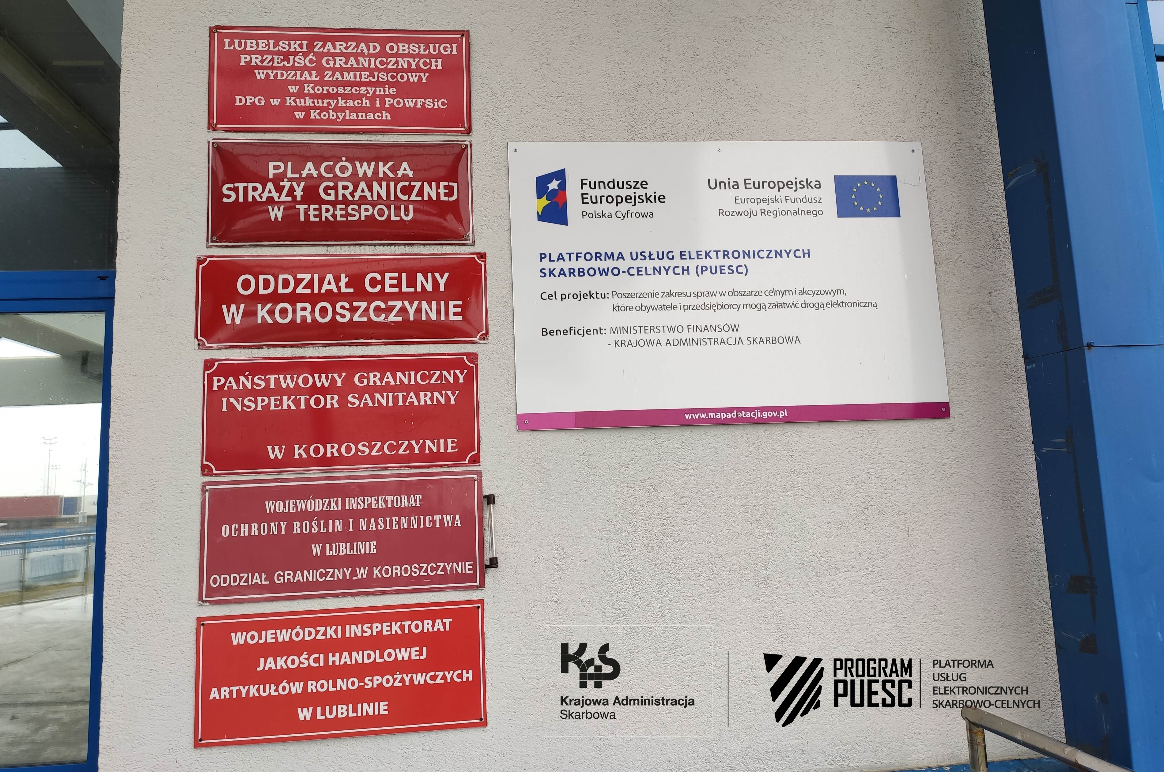 Na środku strony widać ścianę z czerwonymi tabliczkami informującymi o instytucjach mieszczących się w budynku. Na ścianie umieszczone są logotypy: Krajowa Administracja Skarbowa, Fundusze Europejskie Polska Cyfrowa, System Informacyjny Skarbowo-Celny, Ministerstwo Finansów, Unia Europejska Europejski Fundusz Rozwoju Regionalnego. 