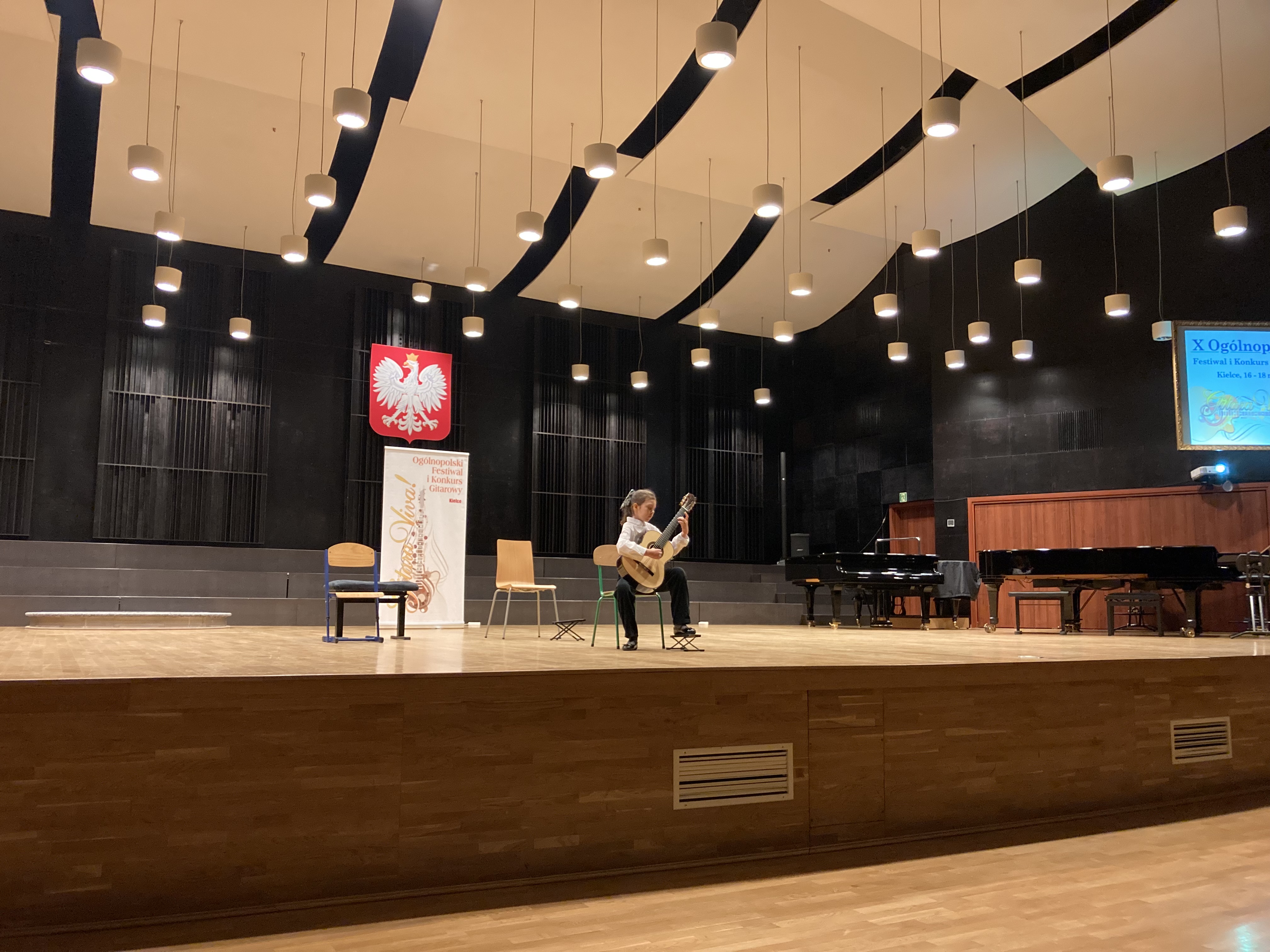 Sala koncertowa Zespołu Szkół Muzycznych w Kielcach, ogromna scena. Na środku sceny występuje uczennica naszej szkoły Lena Torba podczas X Ogólnopolskiego Festiwalu i Konkursu "Gitara Viva" w dniu 16 marca 2023 r. Na środku ściany wisi duże godło Polski, na suficie wisi mnóstwo żyrandoli. Uczennica siedzi na krześle po środku sceny, podczas występu, po jej lewej stronie stoją dwa krzesła, za nią baner z napisem "Gitara Viva!"