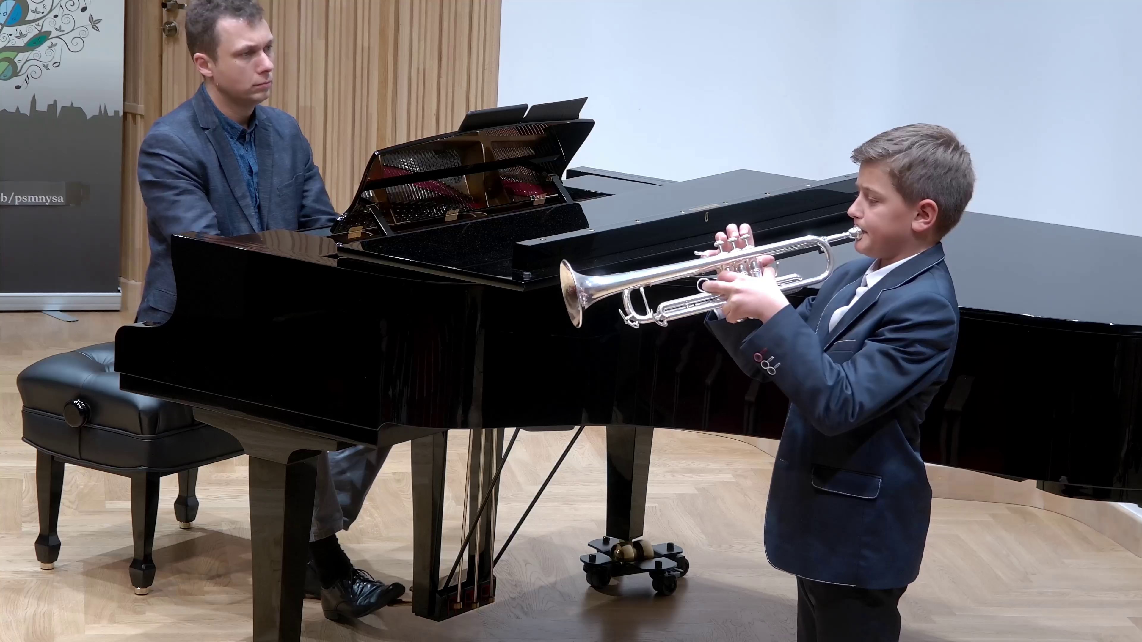 Zdjęcie z koncertu uczniów z klas instrumentów dętych PSM I i II stopnia w Nysie - Sala Koncertowa. Na pierwszym planie trębacz z instrumentem, oraz pianista grający na czarnym fortepianie. W tle baner PSM Nysa.