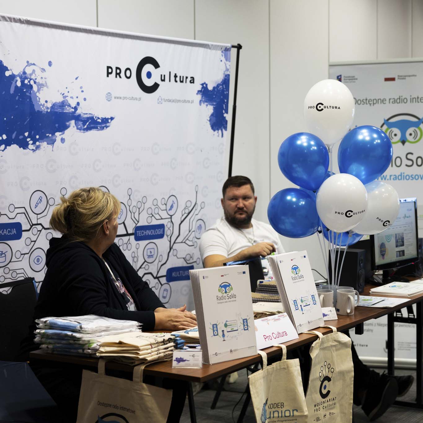 Kobieta i mężczyzna siedzą przy biurku na którym znajdują się materiały reklamowe Fundacji Pro Cultra i Radia SoVo.