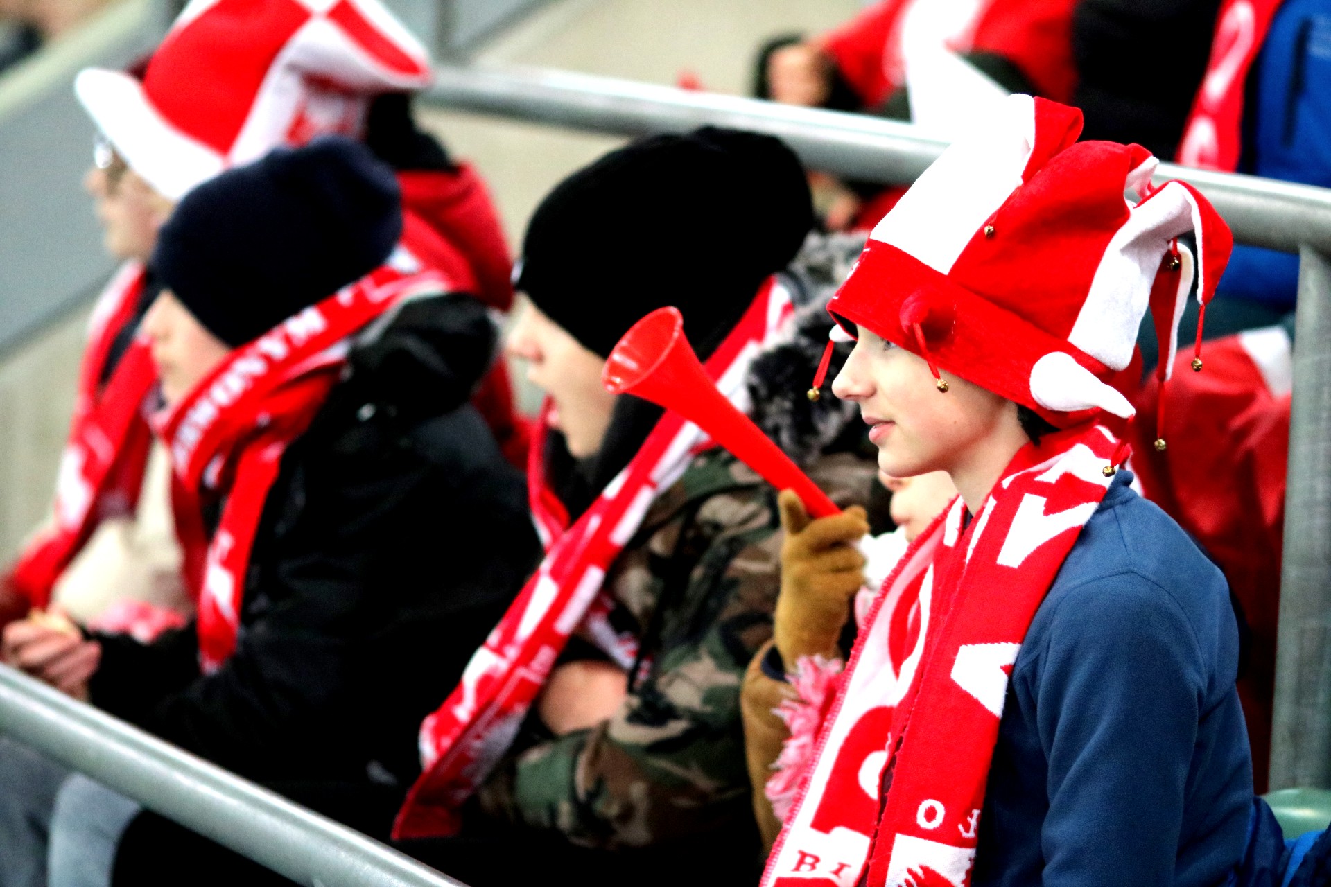 Grupa dzieci na trybunie stadionu, mają biało-czerwone czapki i szaliki, jeden chłopiec trzyma czerwoną trąbkę.
