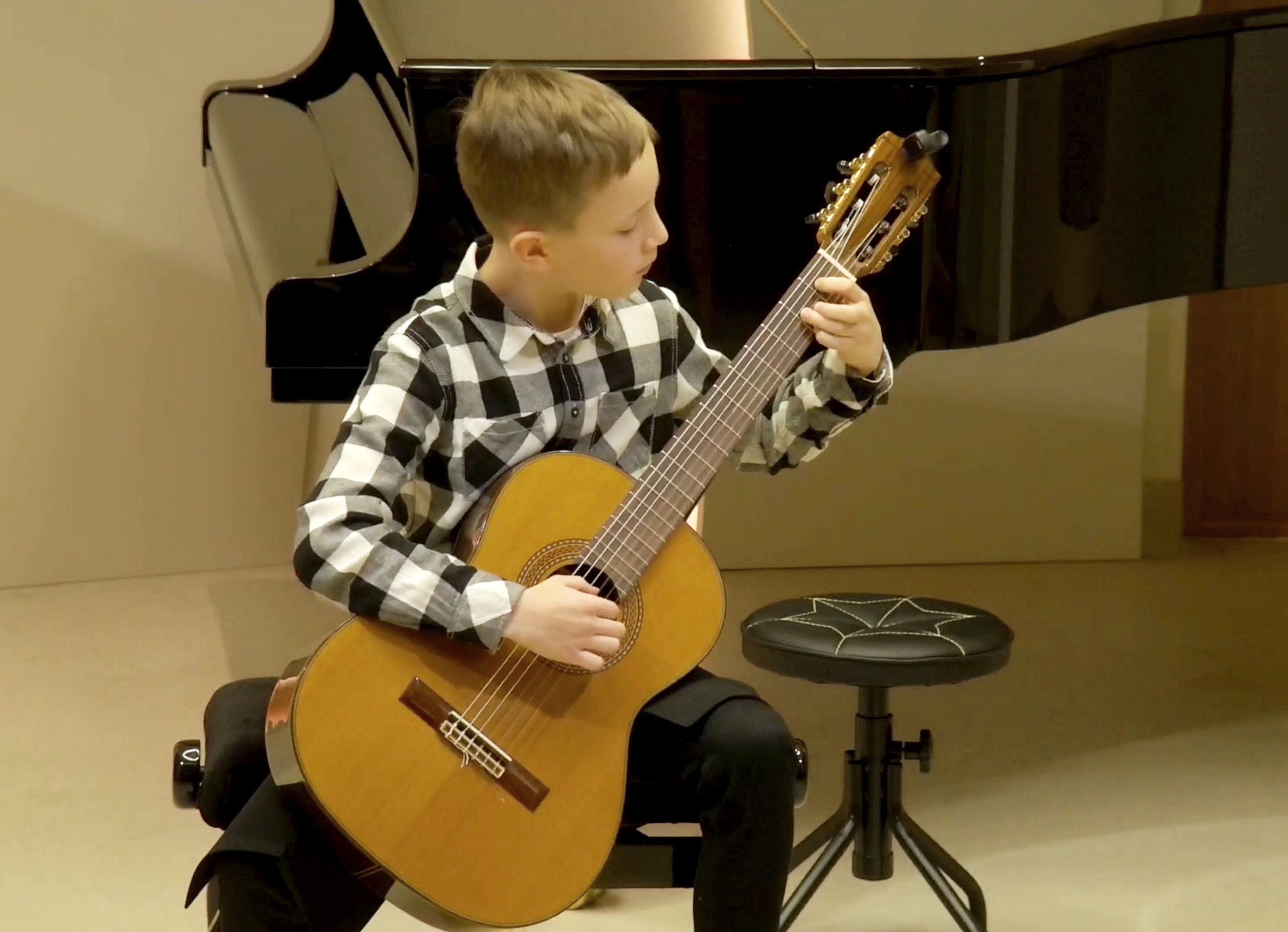 Zdjęcie. Na pierwszym planie młody gitarzysta gra na gitarze. W tle stoi czarny fortepian i niewykorzystywany w tym momencie stołek gitarowy. Koncert odbywa się w Sali Kameralnej PSM w Nysie.