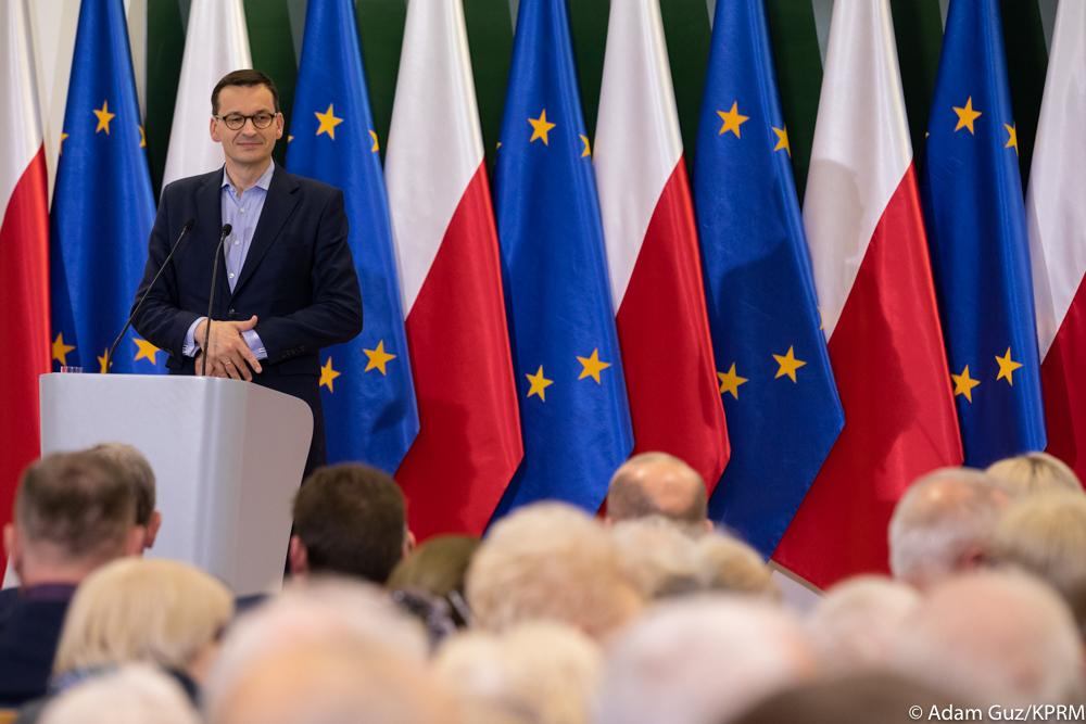 Premier Mateusz Morawiecki przemawia na tle flag Polski i Unii Europejskiej.