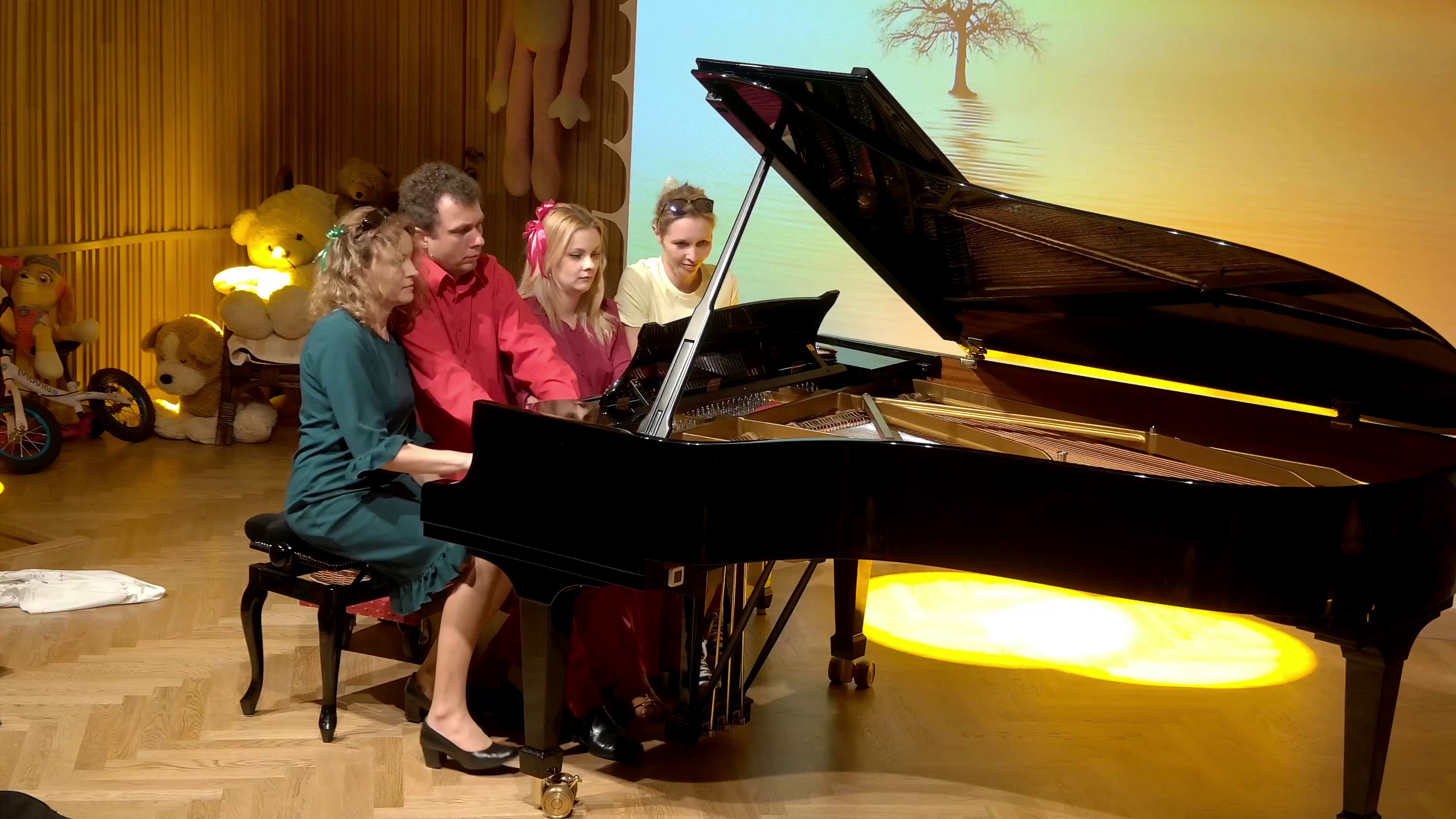 Zdjęcie. na estradzie sali koncertowej występuje czworo dorosłych pianistów. Graja na jednym dużym czarnym otwartym fortepianie. W tle różne zabawki ułożone pod ścianą.