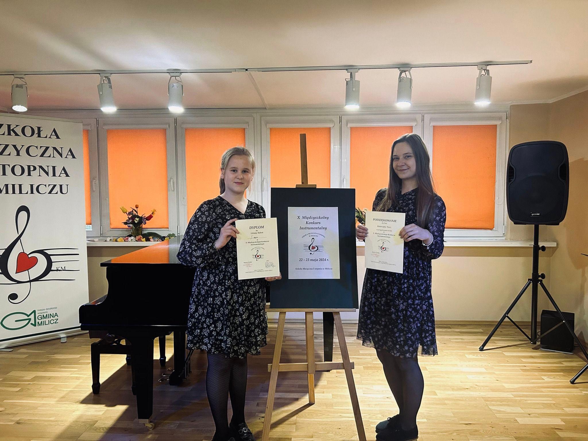 Na zdjęciu widoczna jest uczennica wraz z nauczycielką skrzypiec. Obie trzymają w rękach dyplomy konkursowe. w tle widoczny jest fortepian oraz baner konkursu.