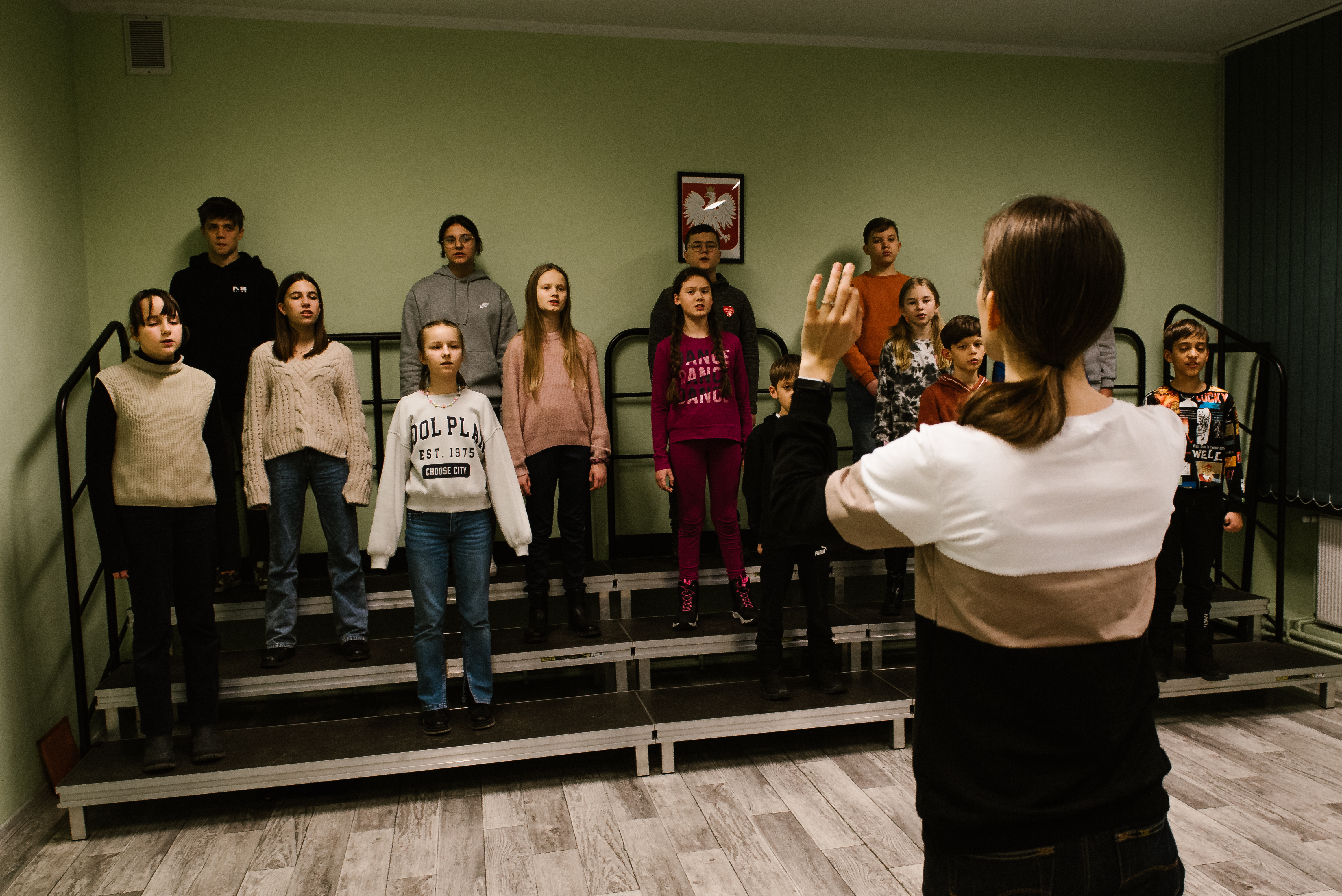 Na zdjęciu znajduje się nauczycielka Aleksandra Szymańska oraz grupa uczniów. Nauczycielka stoi przodem do dzieci z uniesionymi rękami, dyrygując śpiewającym uczniom stojącym na podeście. Na ścianie wisi godło polski. 