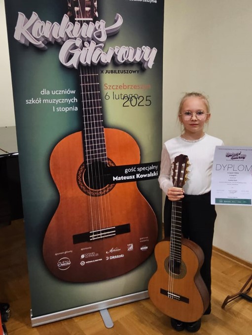 Zdjęcie Zuzanny Stach na konkursie w Szczebrzeszynie. Uczennica z gitarą w ręce, w tle baner konkursowy.