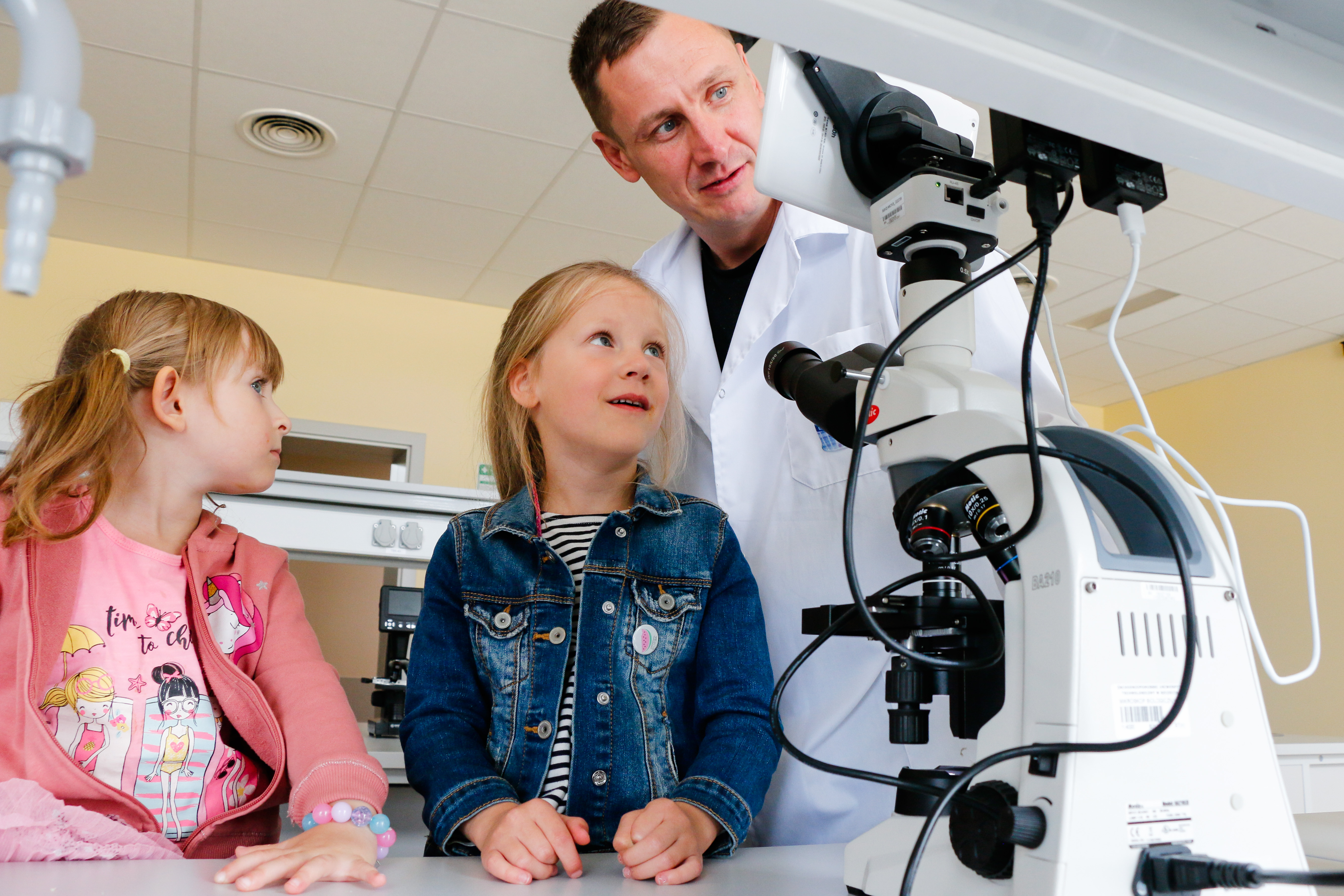 Laboratorium. Młody ciemnowłosy mężczyzna w białym kitlu pokazuje z uśmiechem mikroskop dwóm 7-8-letnim dziewczynkom 